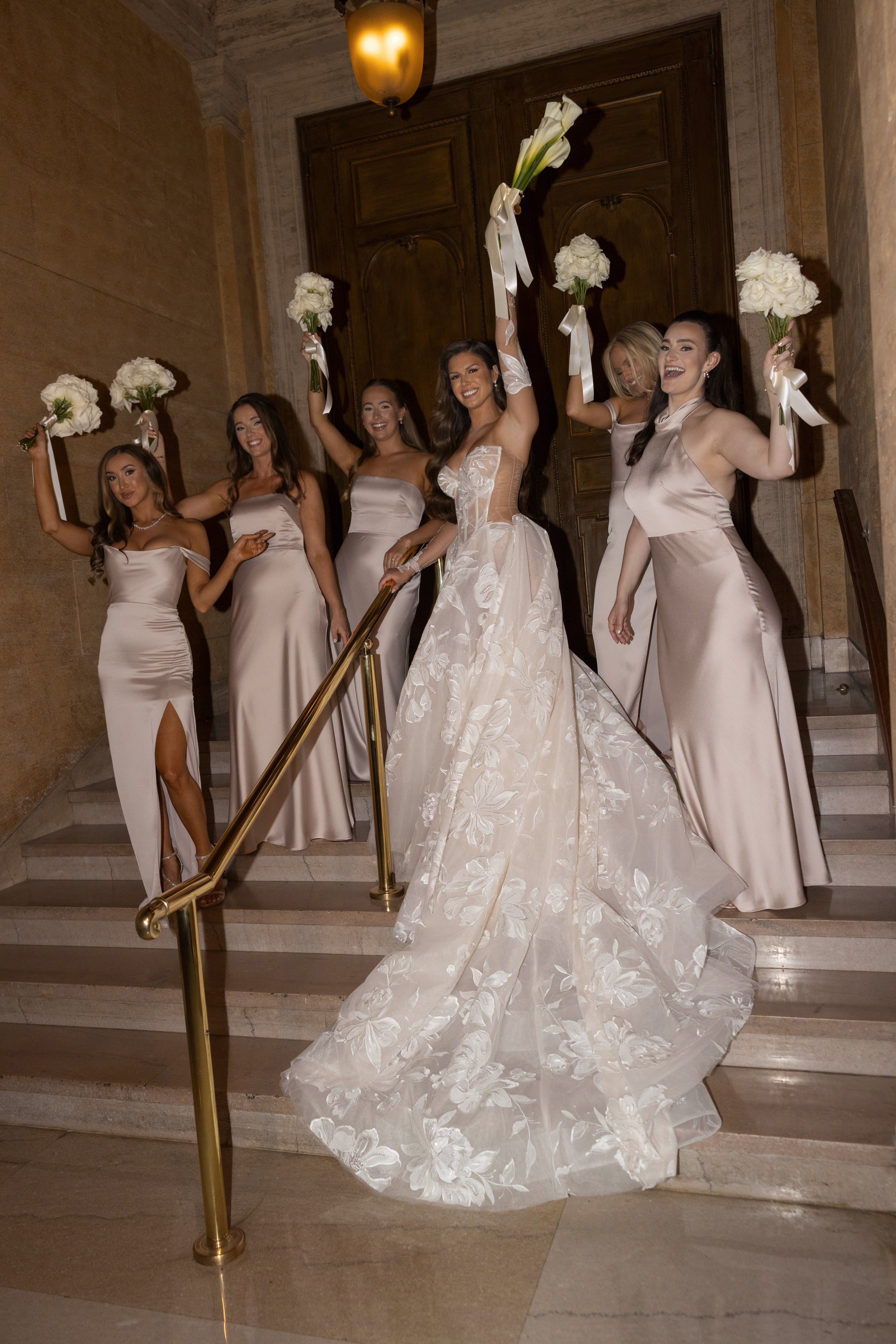 Bride and bridesmaids in white dresses standing on a staircase holding flowers.