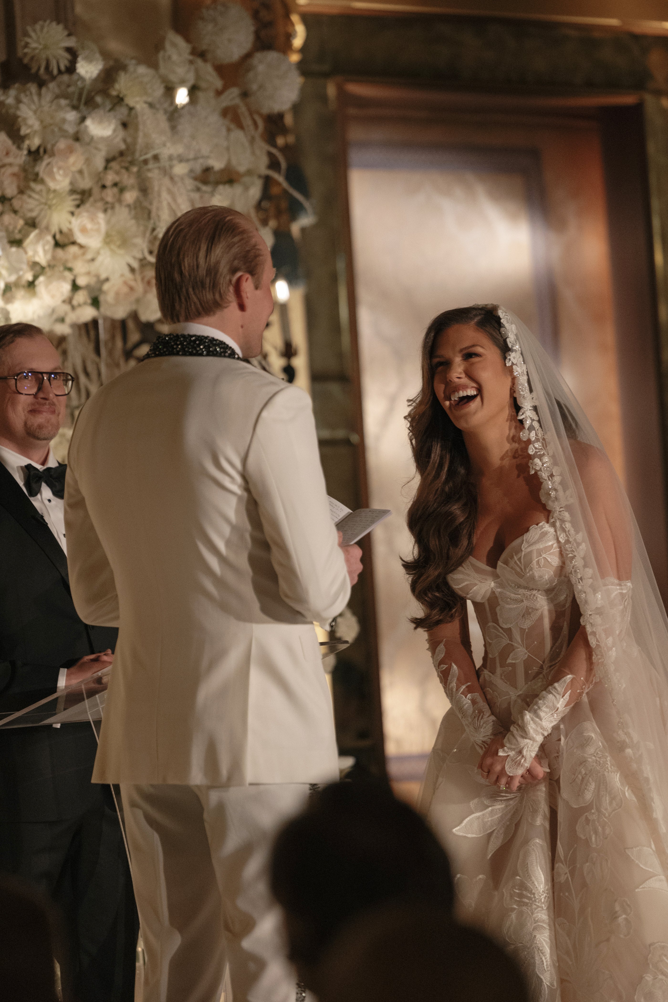 Man in a suit and woman in a wedding dress standing in front of a mirror with floral decorations.