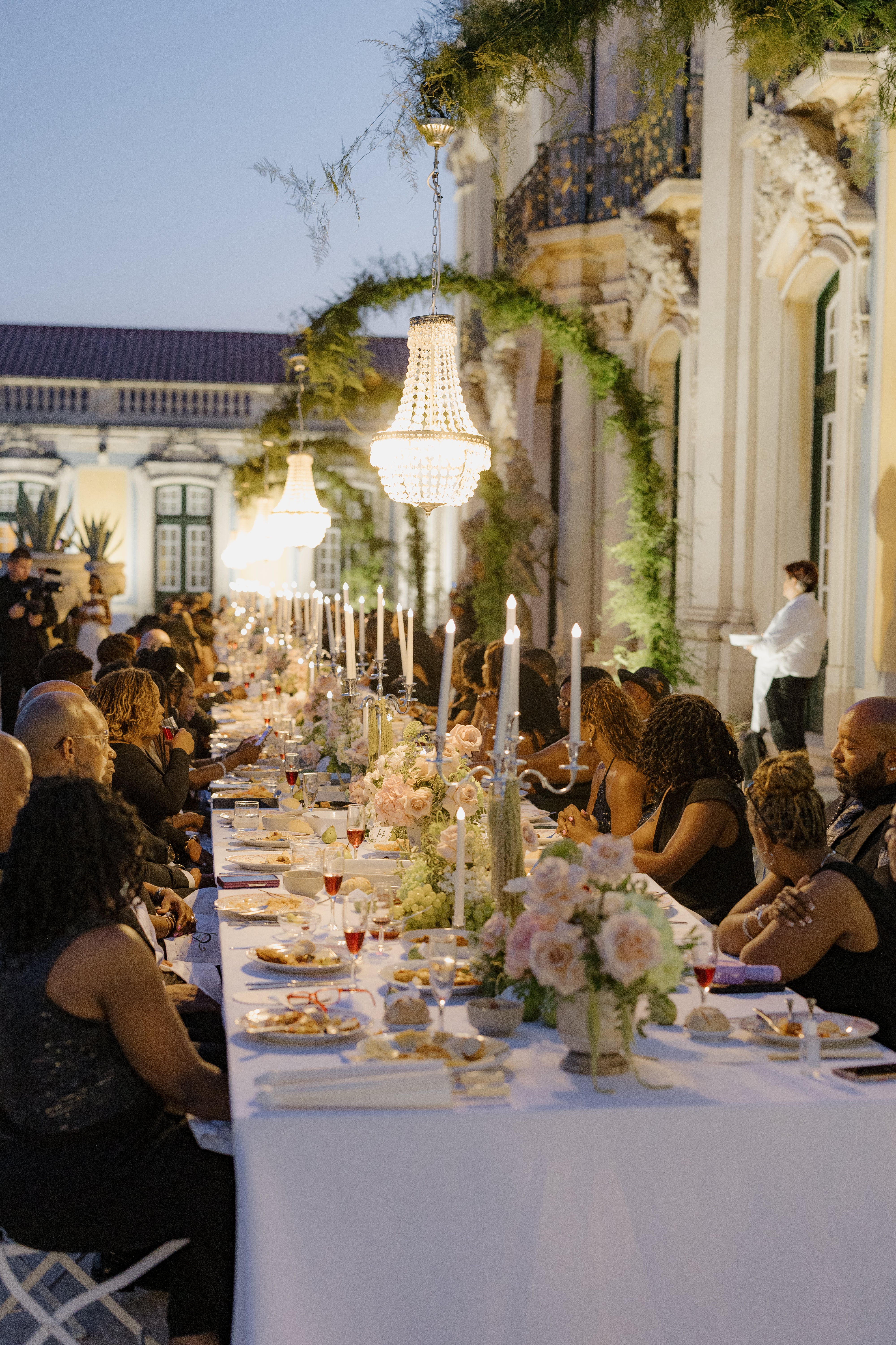 Elegant outdoor dinner event with guests seated at a long table adorned with flowers and candles, set against an architectural backdrop. 