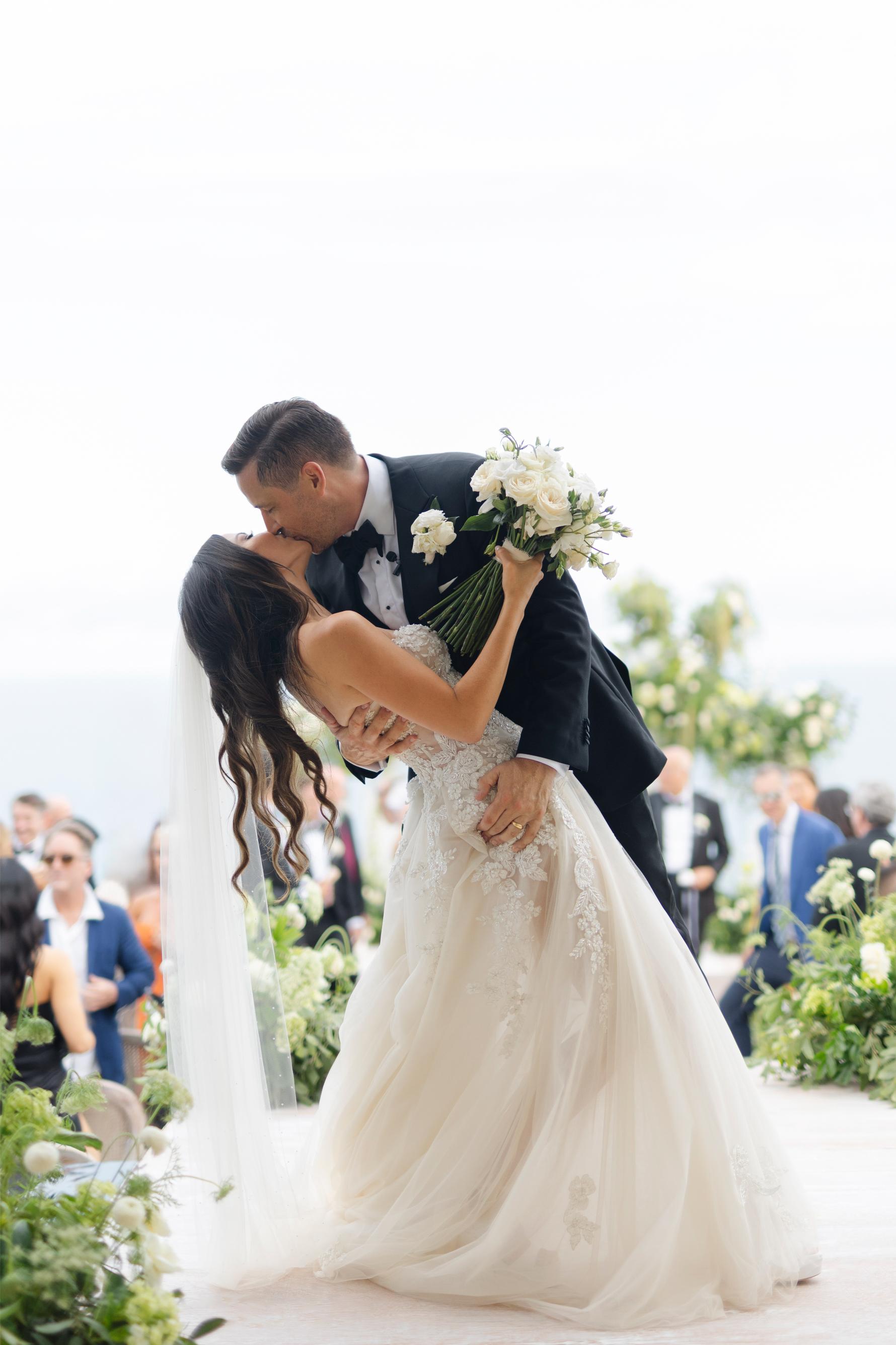 Couple sharing a kiss on their wedding day with a blurred background of guests.