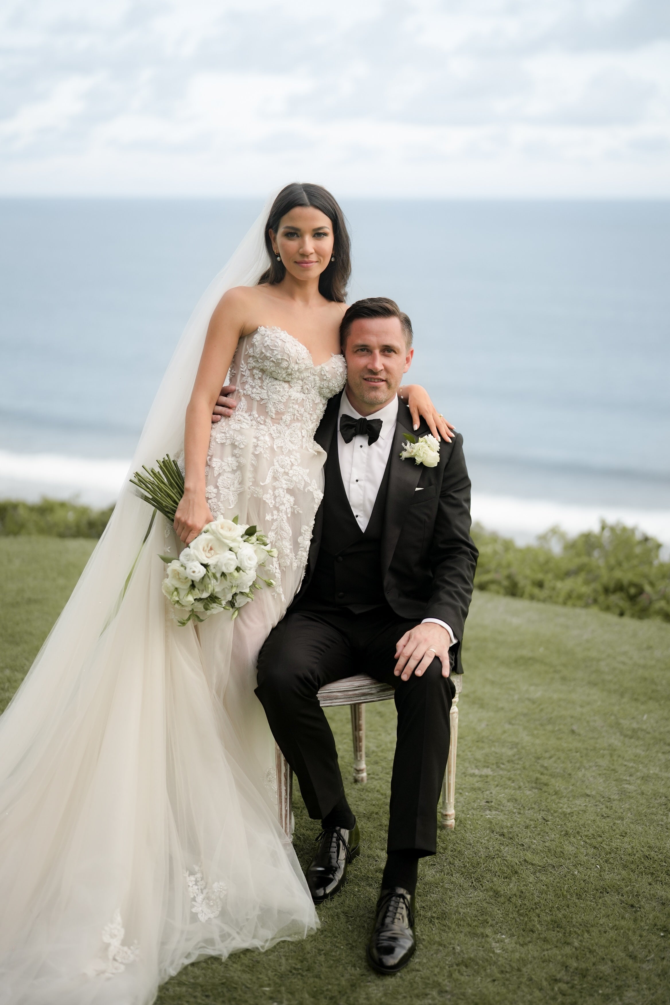 Couple in wedding attire sitting on a grassy area with ocean view