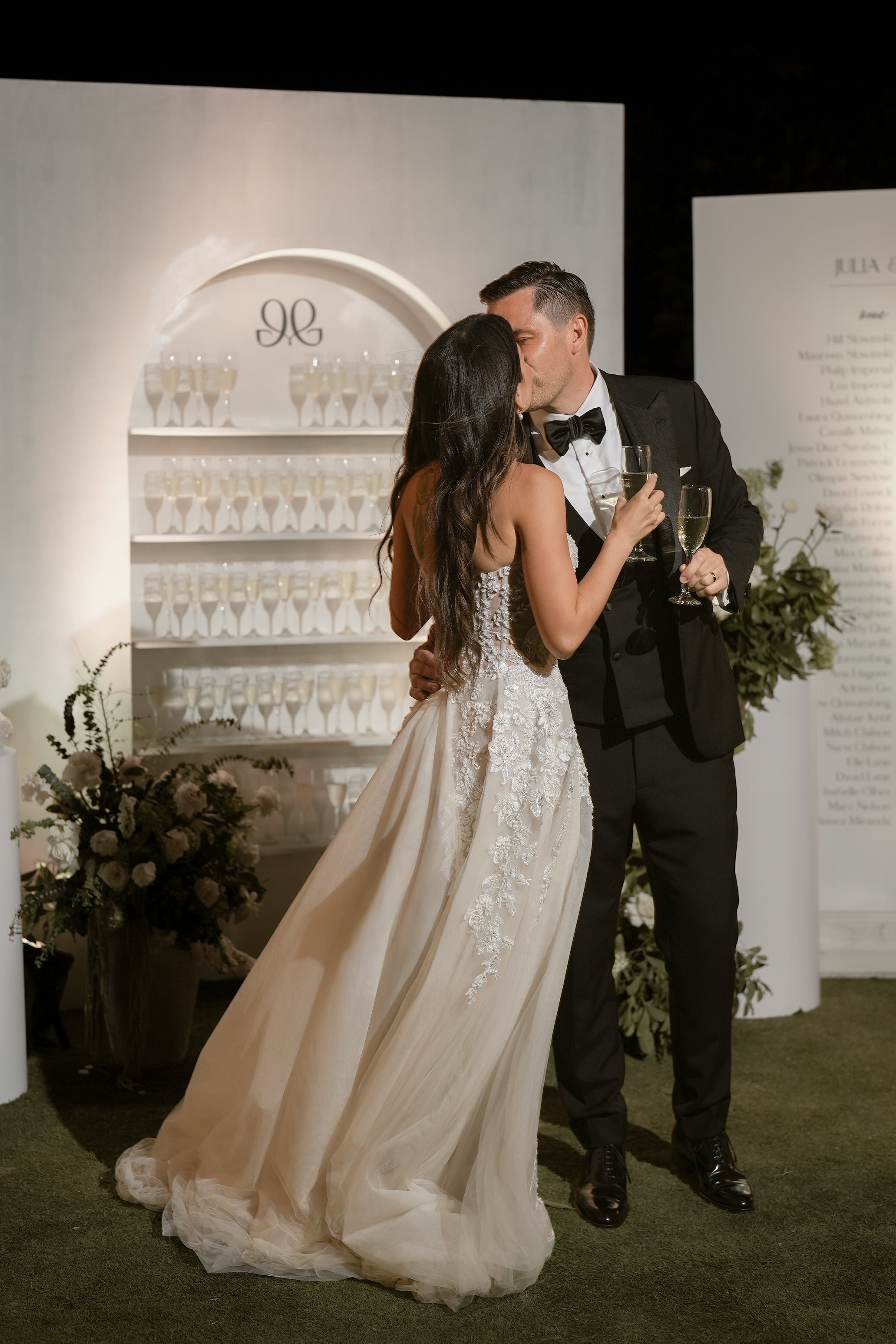 Man and woman in formal attire embracing with champagne glasses against a decorative backdrop.