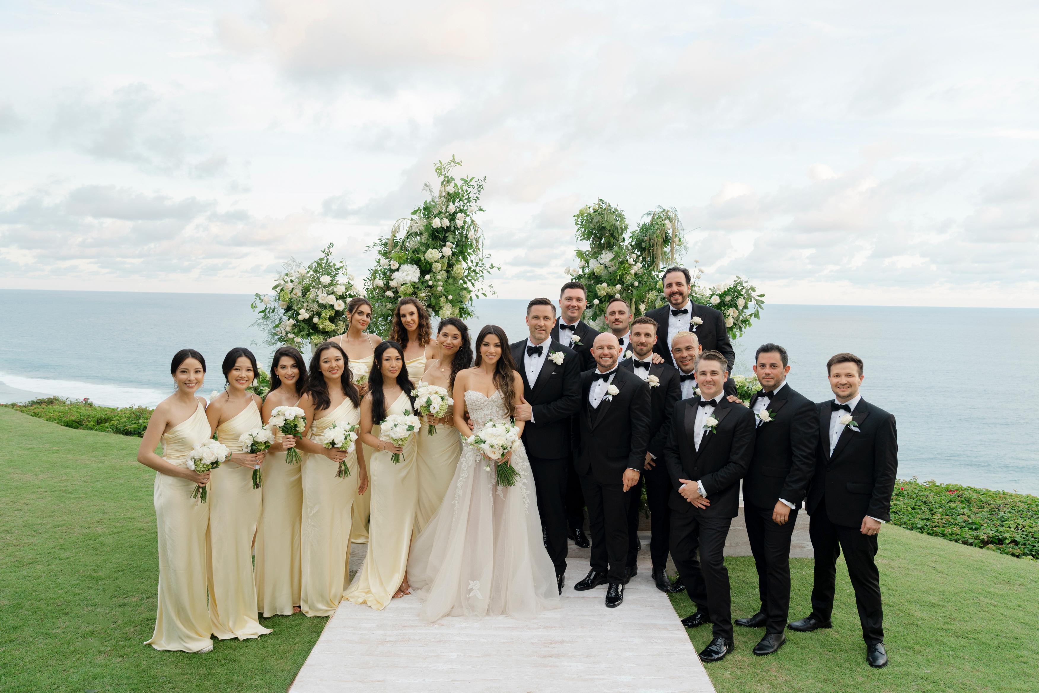 Wedding party posing on a grassy area with floral decorations and ocean view.