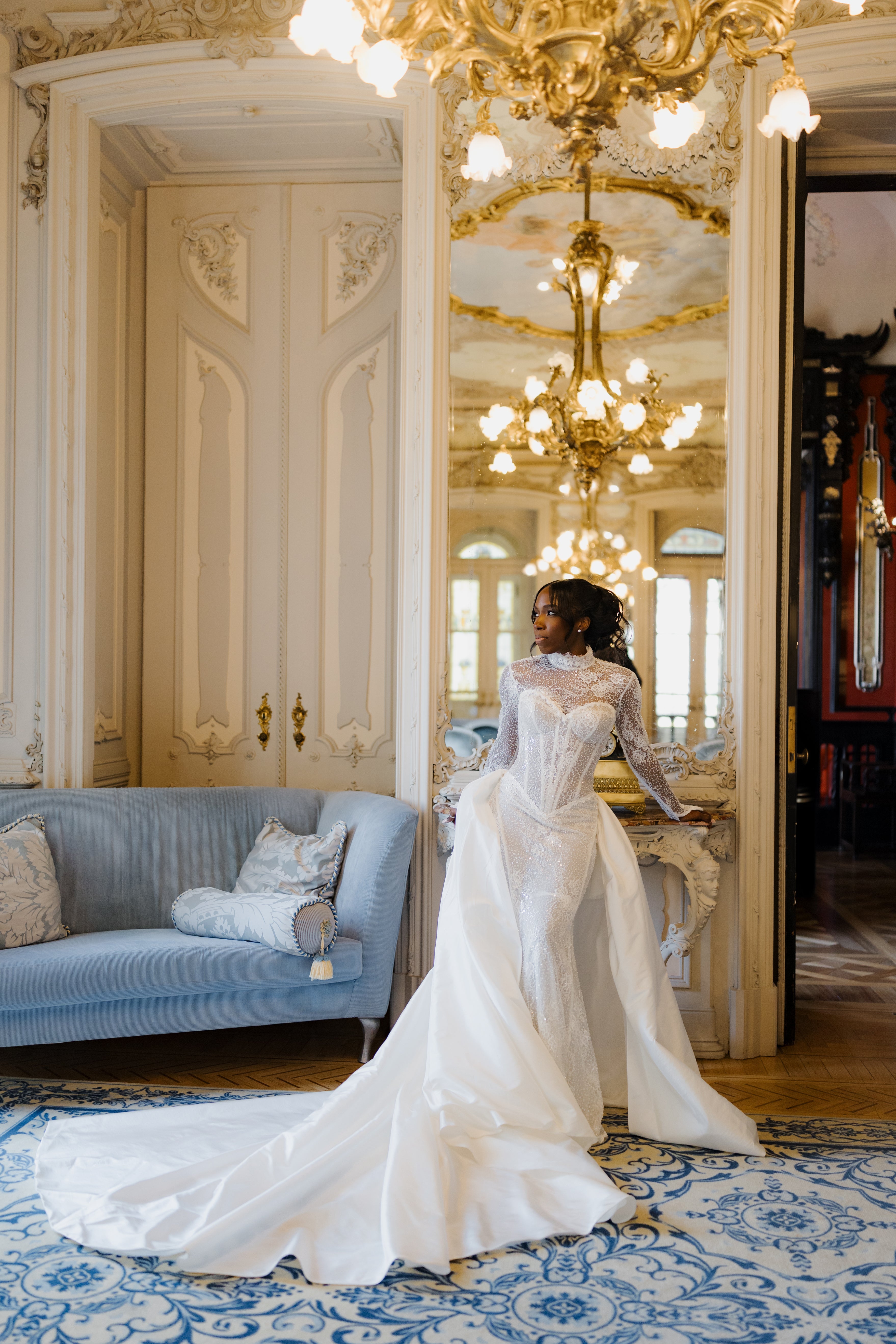 Woman in a white wedding dress standing in an elegant room with chandeliers and a blue sofa.