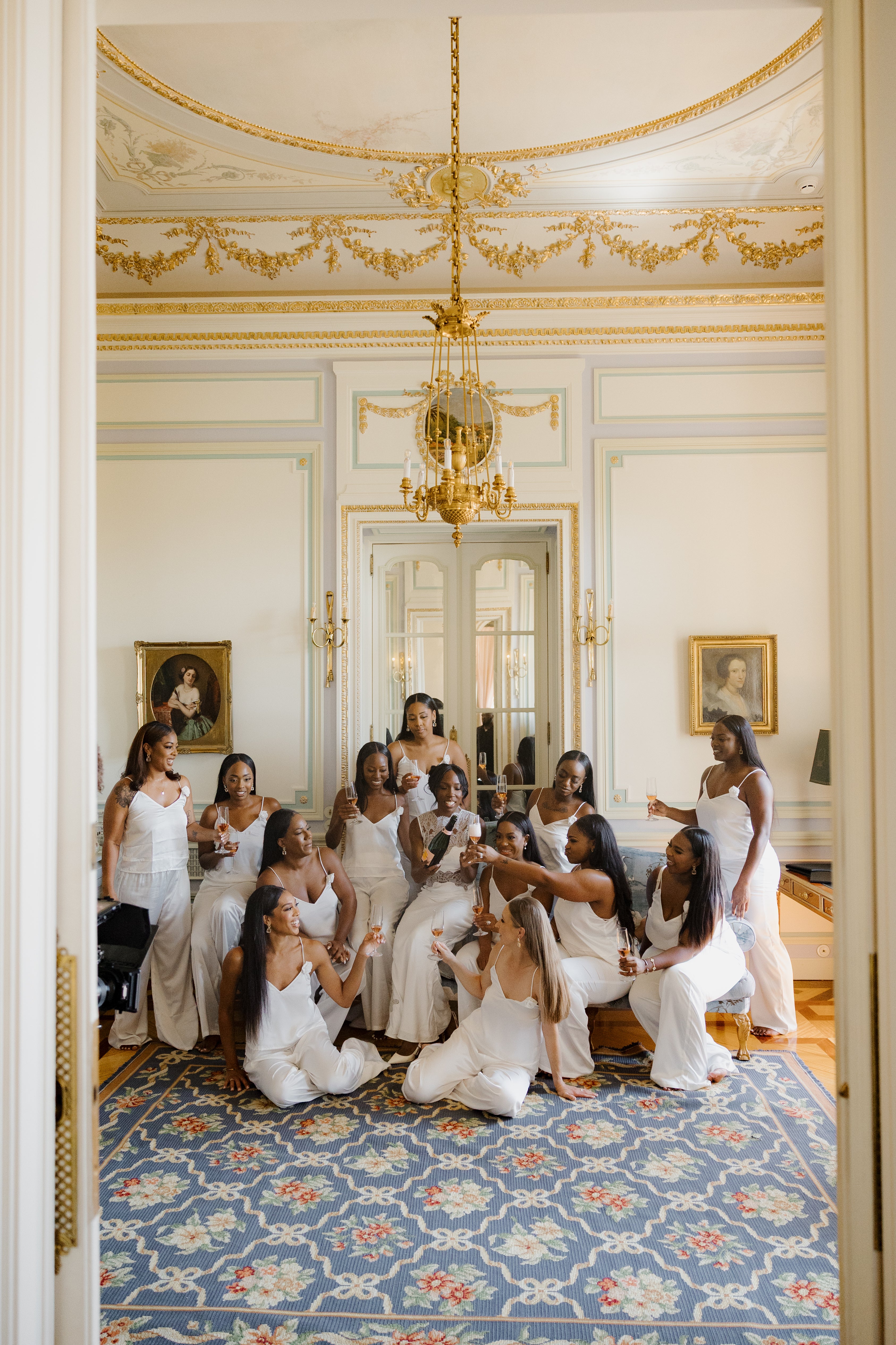 Group of women in formal attire in a grand room with decorative ceiling and chandelier.