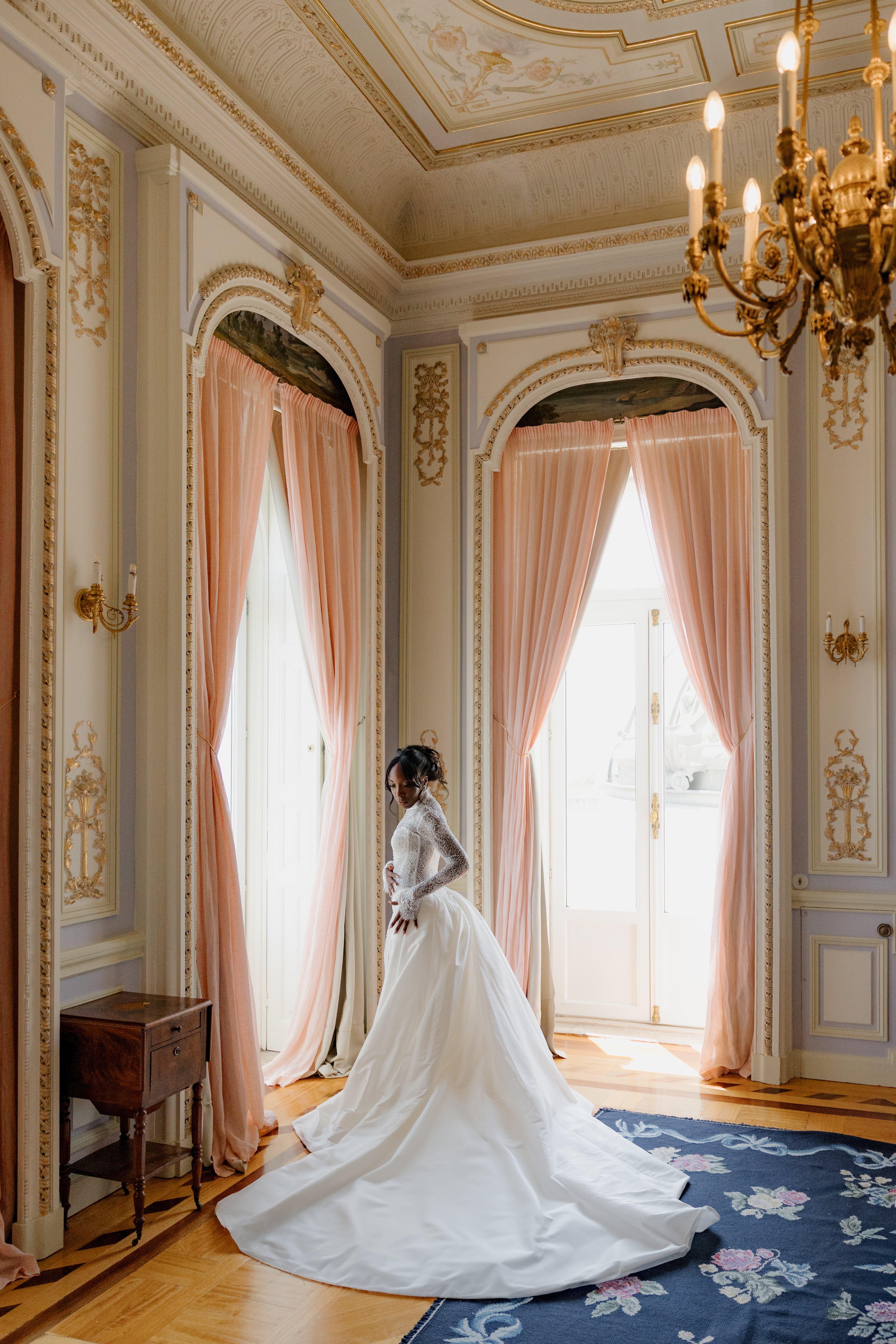 Woman in a white wedding dress standing in an opulent room with pink curtains and a chandelier.