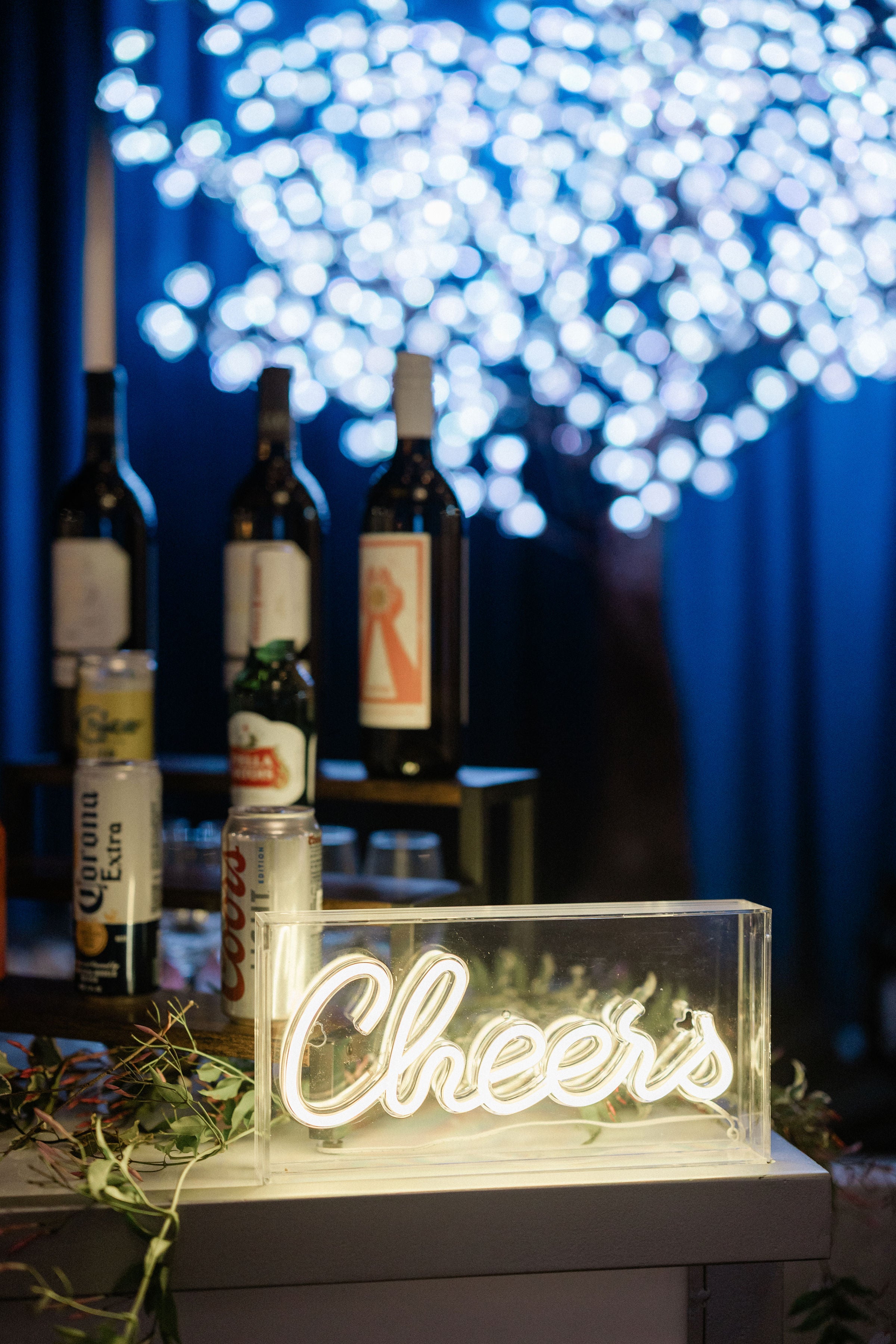 Neon 'Cheers' sign with bottles in the background under string lights