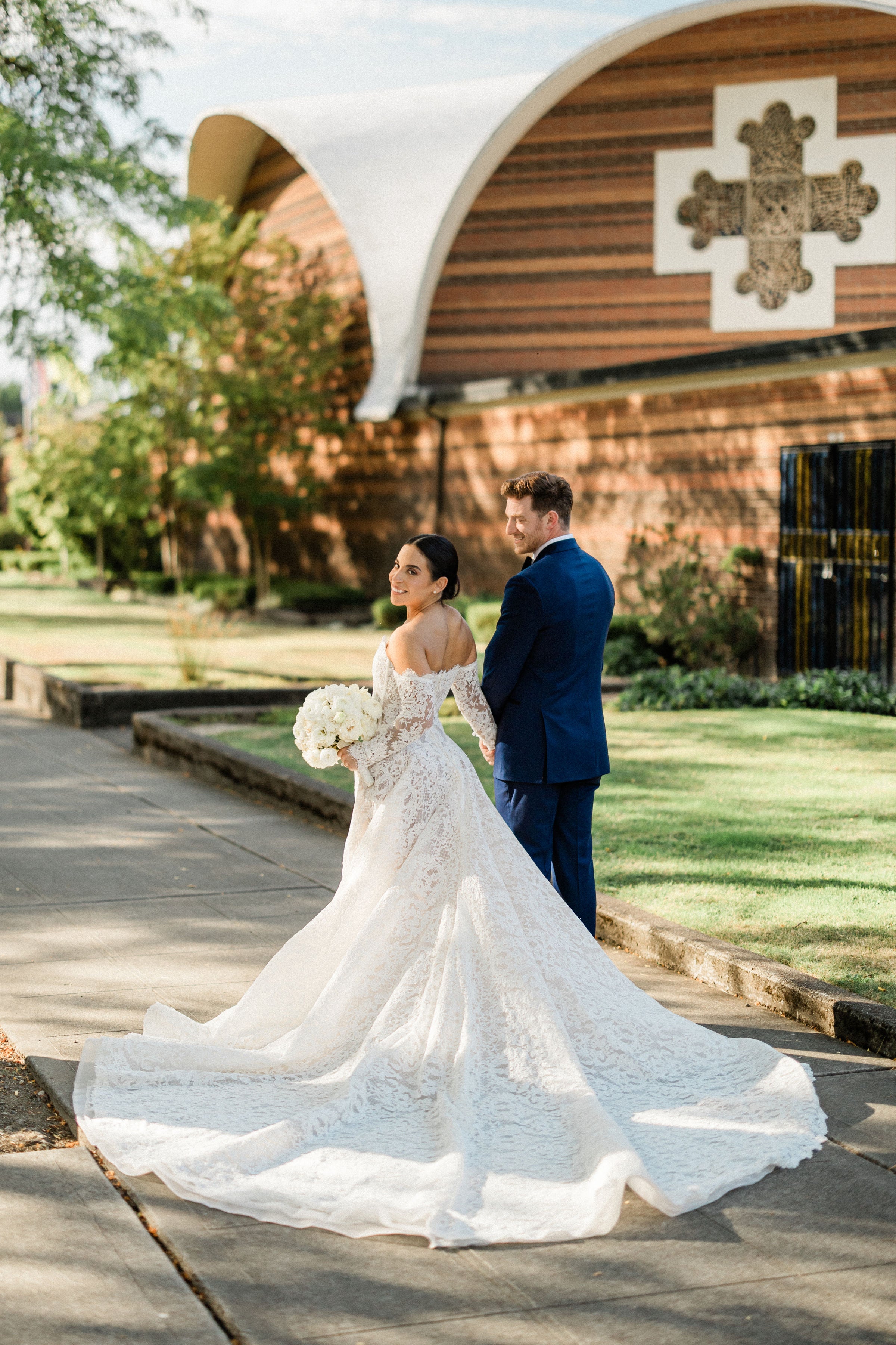 GL Bride Megan wearing Galia Lahav Splendid with her husband,  standing in front of a church with a cross on the wall.