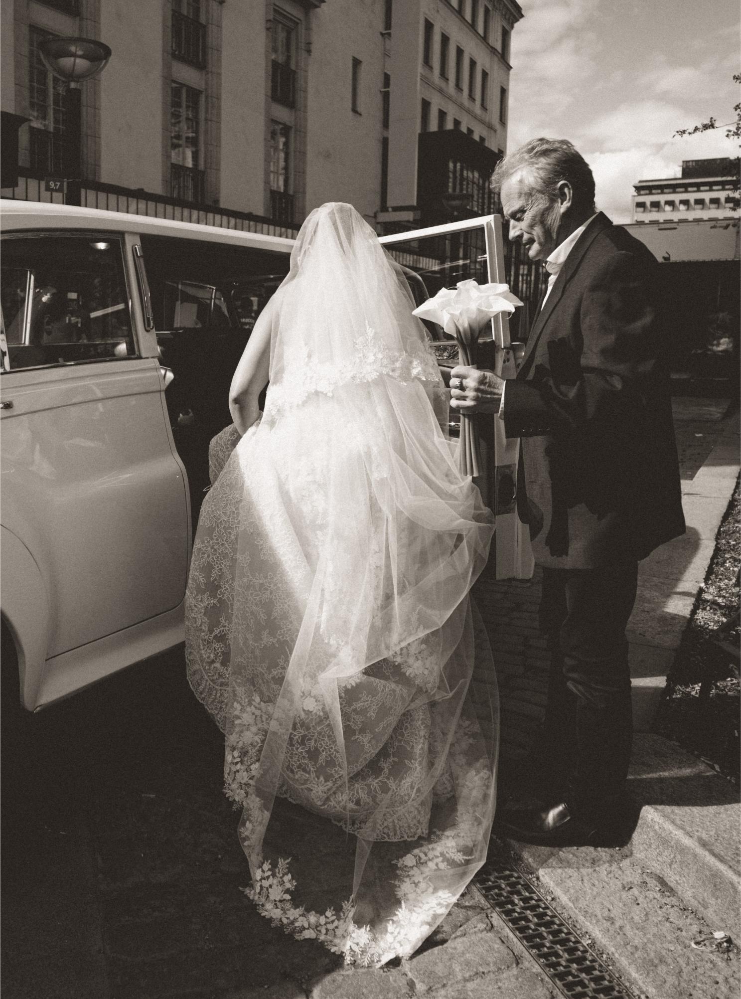 Black and white photograph of a bride being helped out of a car by a man.