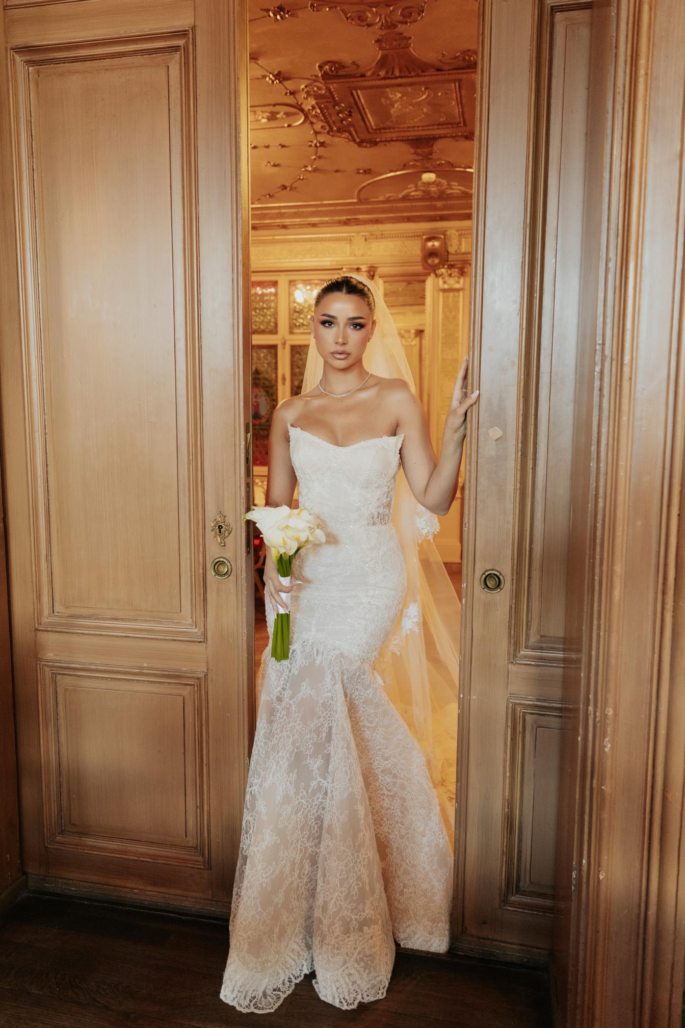 GL Bride Rebecca wearing our Scarlett wedding dress standing in a doorway with a wooden interior.