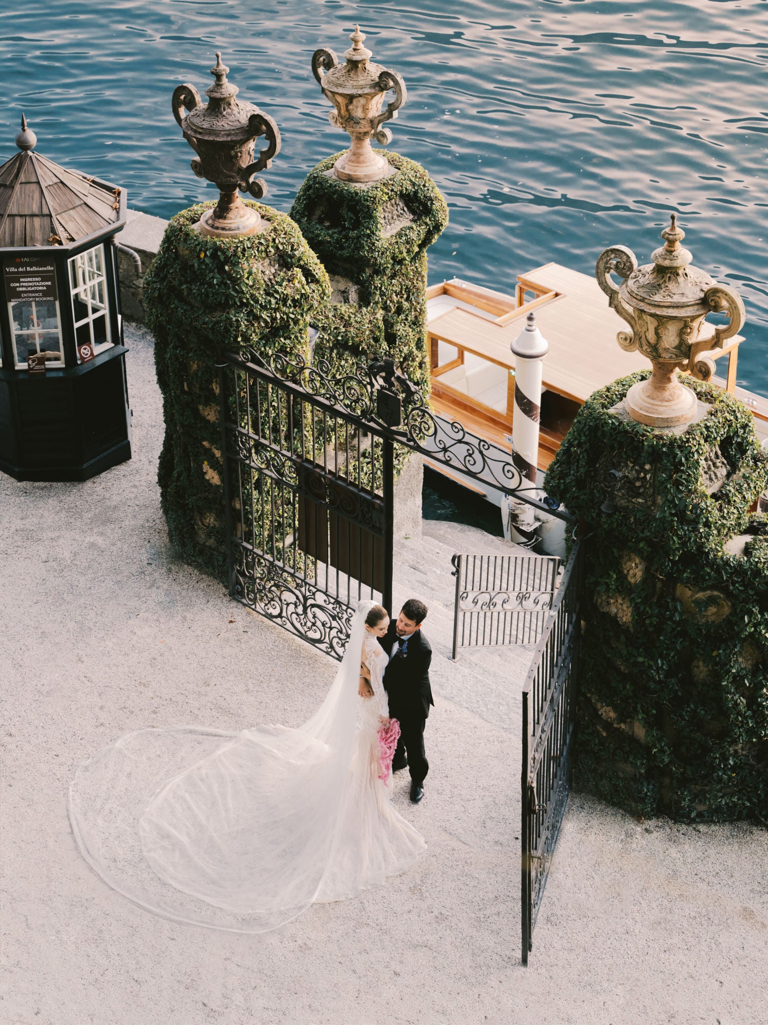 GL Bride Sarah Ingle in our Élysée and her groom walking by ornate gate with decorative details on a waterfront.