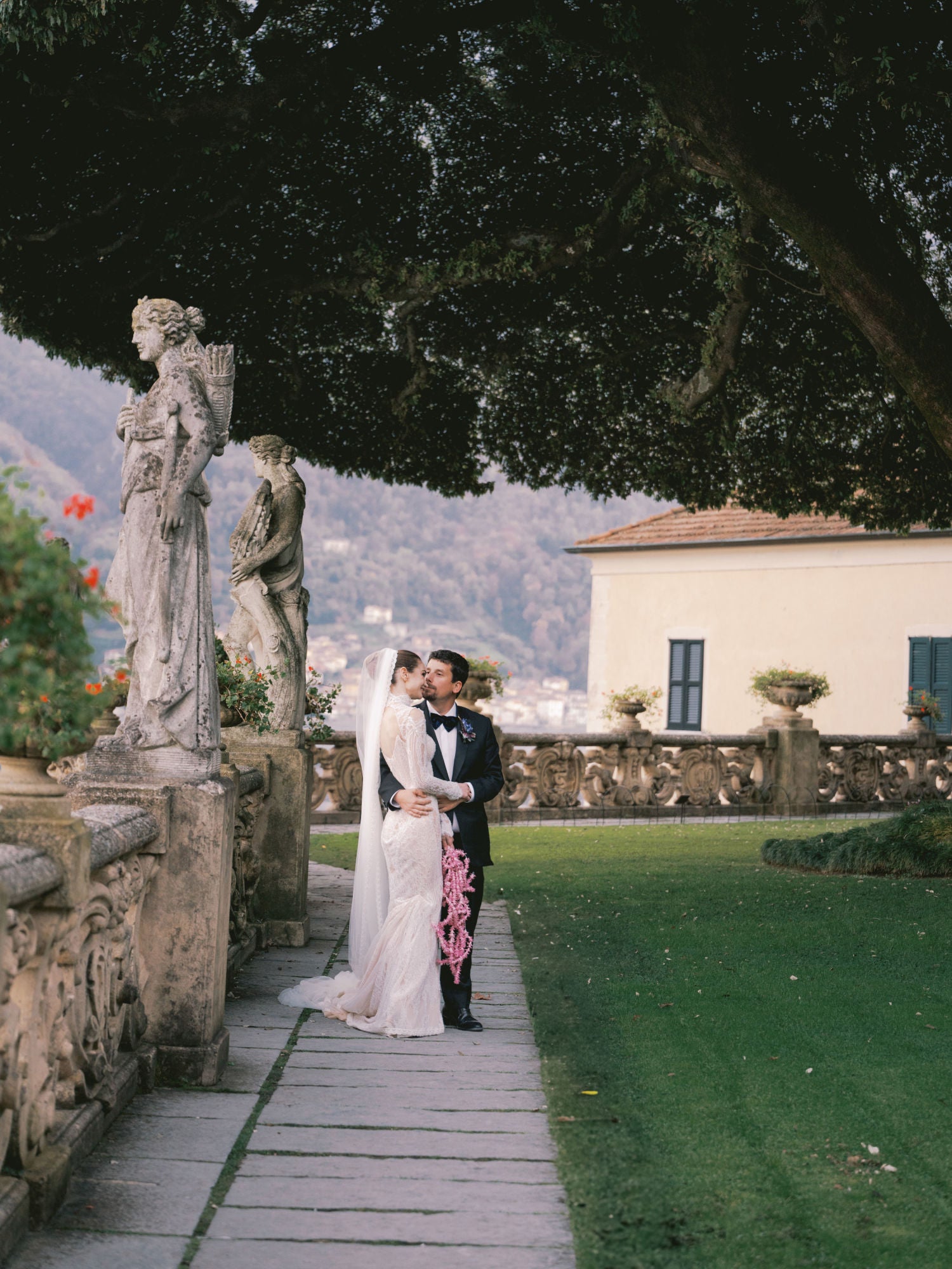 GL Bride Sarah Ingle wearing Galia Lahav's Élysée with her groom embracing on a stone pathway with statues and a scenic background