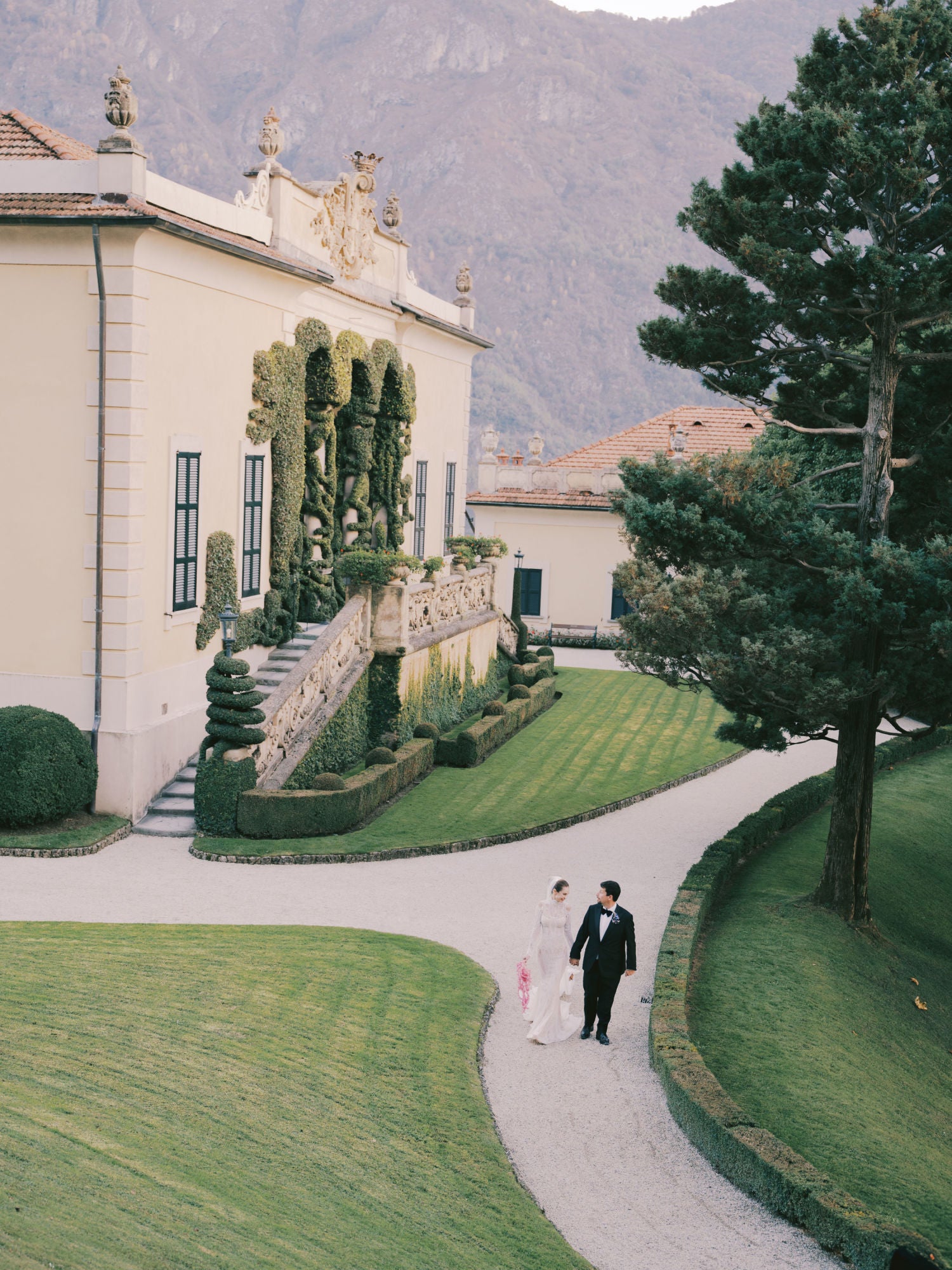 GL Bride Sarah in our Élysée with her husband on a path in front of an elegant building with manicured gardens and mountains in the background.