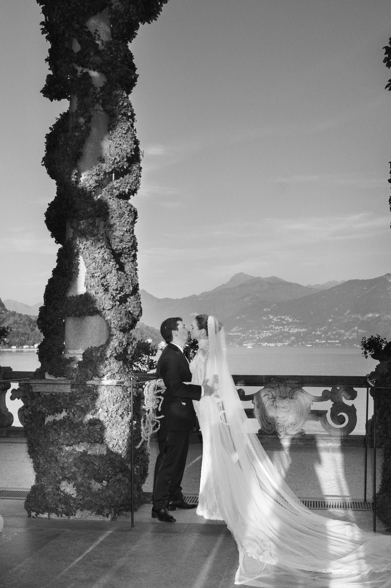 Black and white photo of Sarah Ingle and groom kissing on a balcony with scenic background