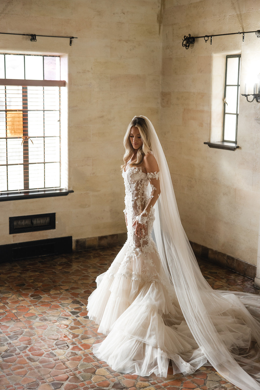 GL Bride Summer in Galia Lahav's Veronica Gown with a long veil standing in a room with stone walls and large windows.
