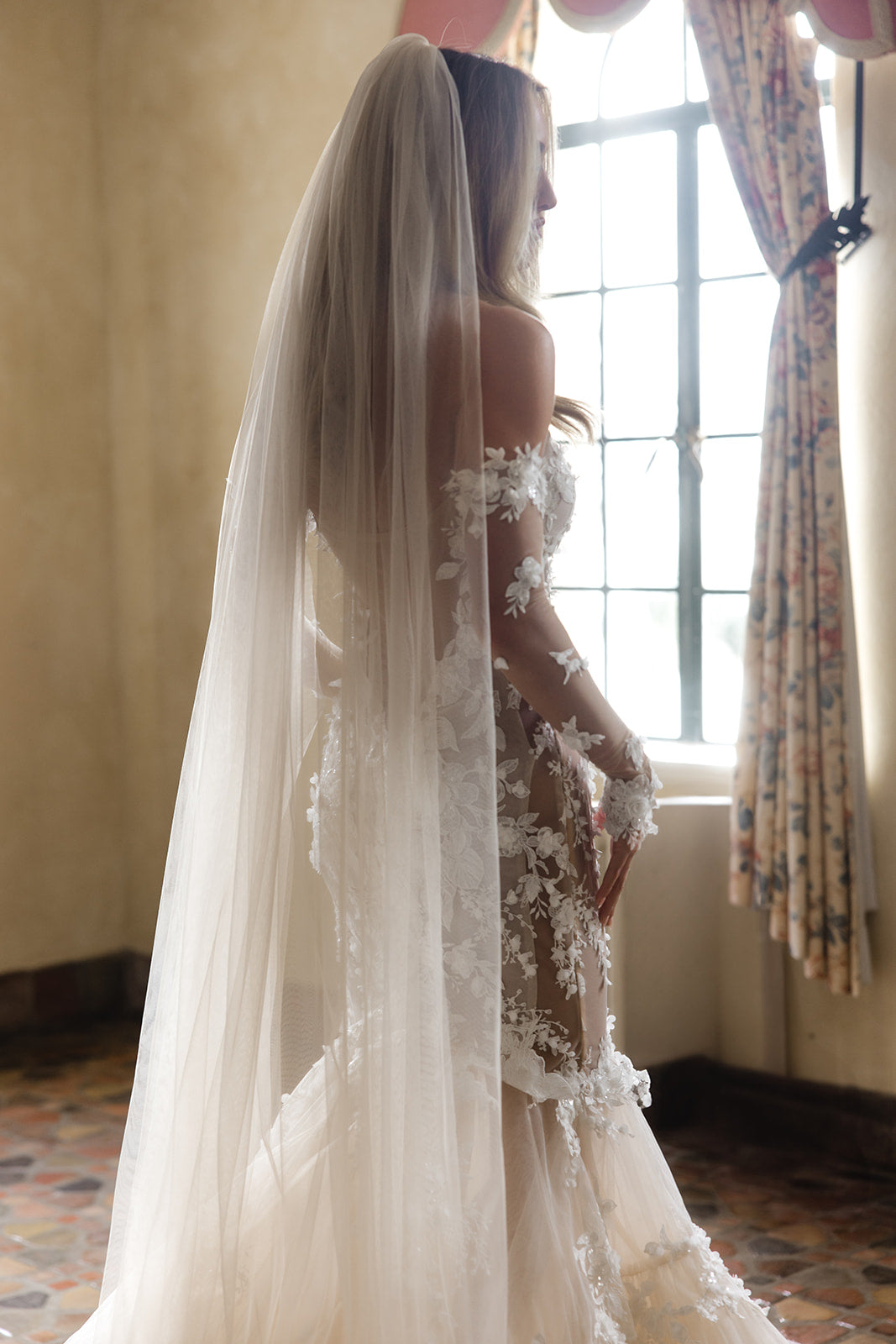 GL Bride Summer in Veronica gown designed by Galia Lahav, with a long veil standing in a room with a window and floral curtains.