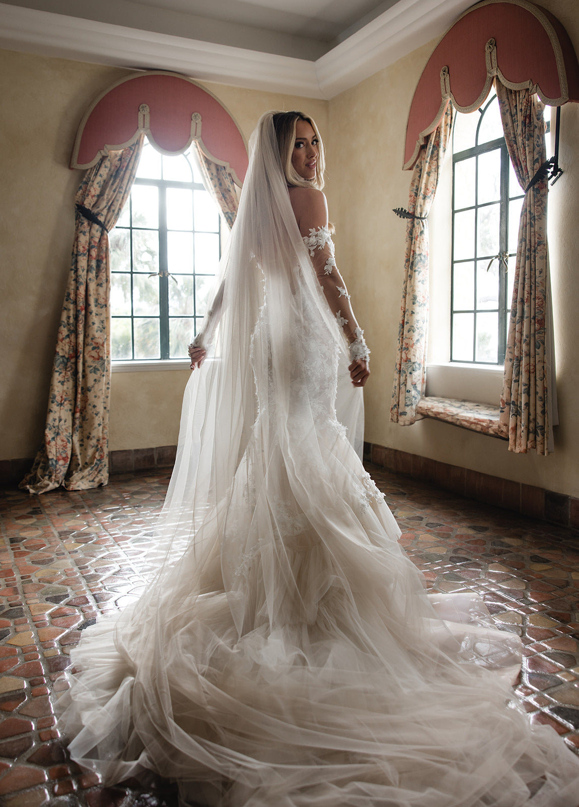 GL Bride Summer in the Veronica Gown with a long veil standing in a room with floral curtains.