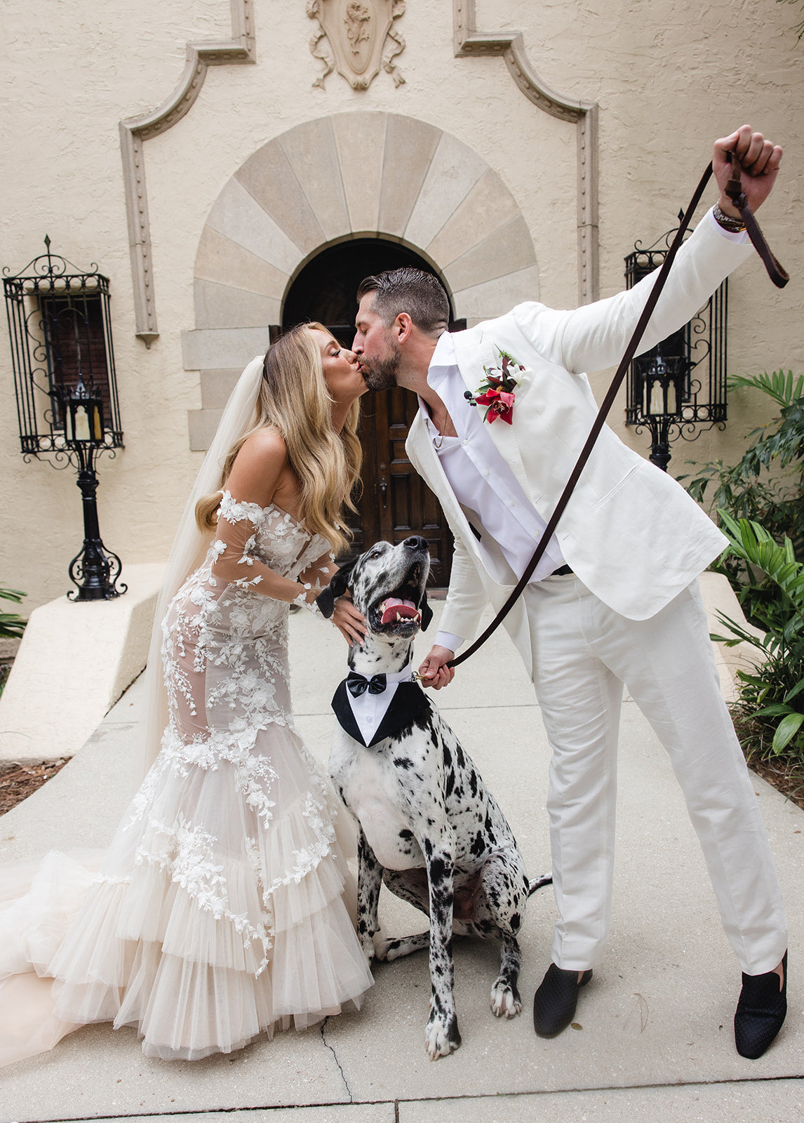 Summer Fox and her husband with a Great Dane dog in front of an elegant building. GL Bride, Summer Fox, wearing the Veronica Gown by Galia Lahav.