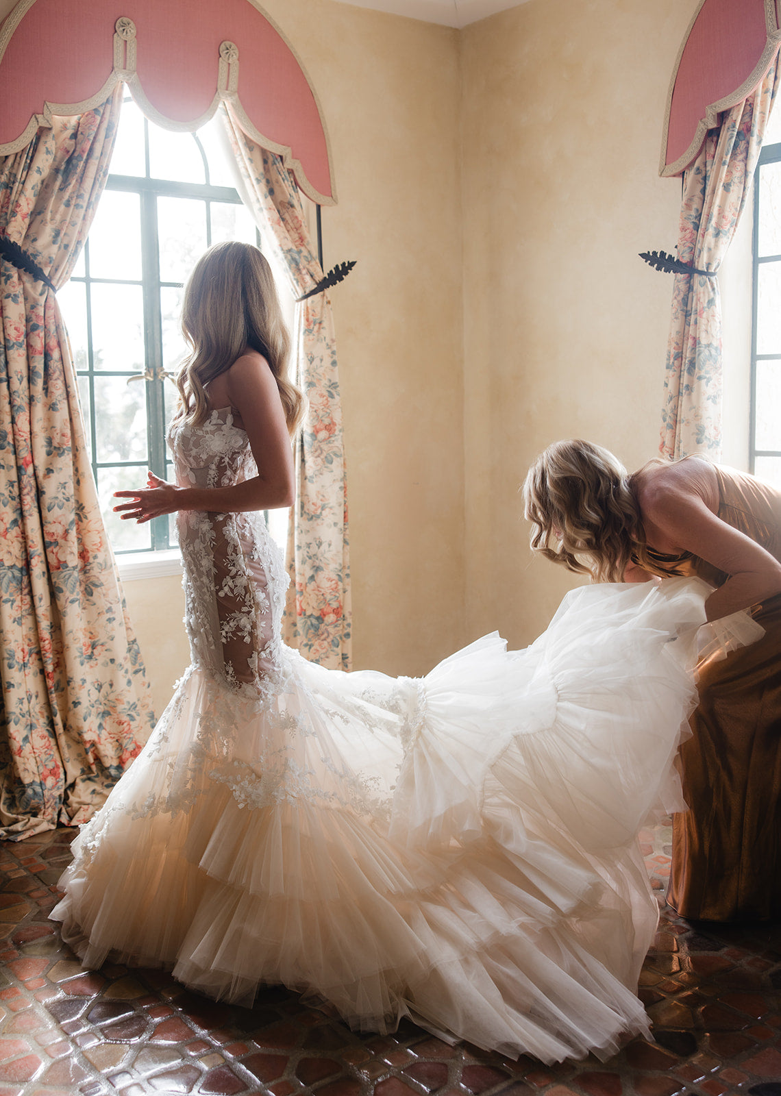 Woman adjusting the train of the GL Bride Summer Fox who wore the lace Veronica dress in a room with floral curtains and a window.