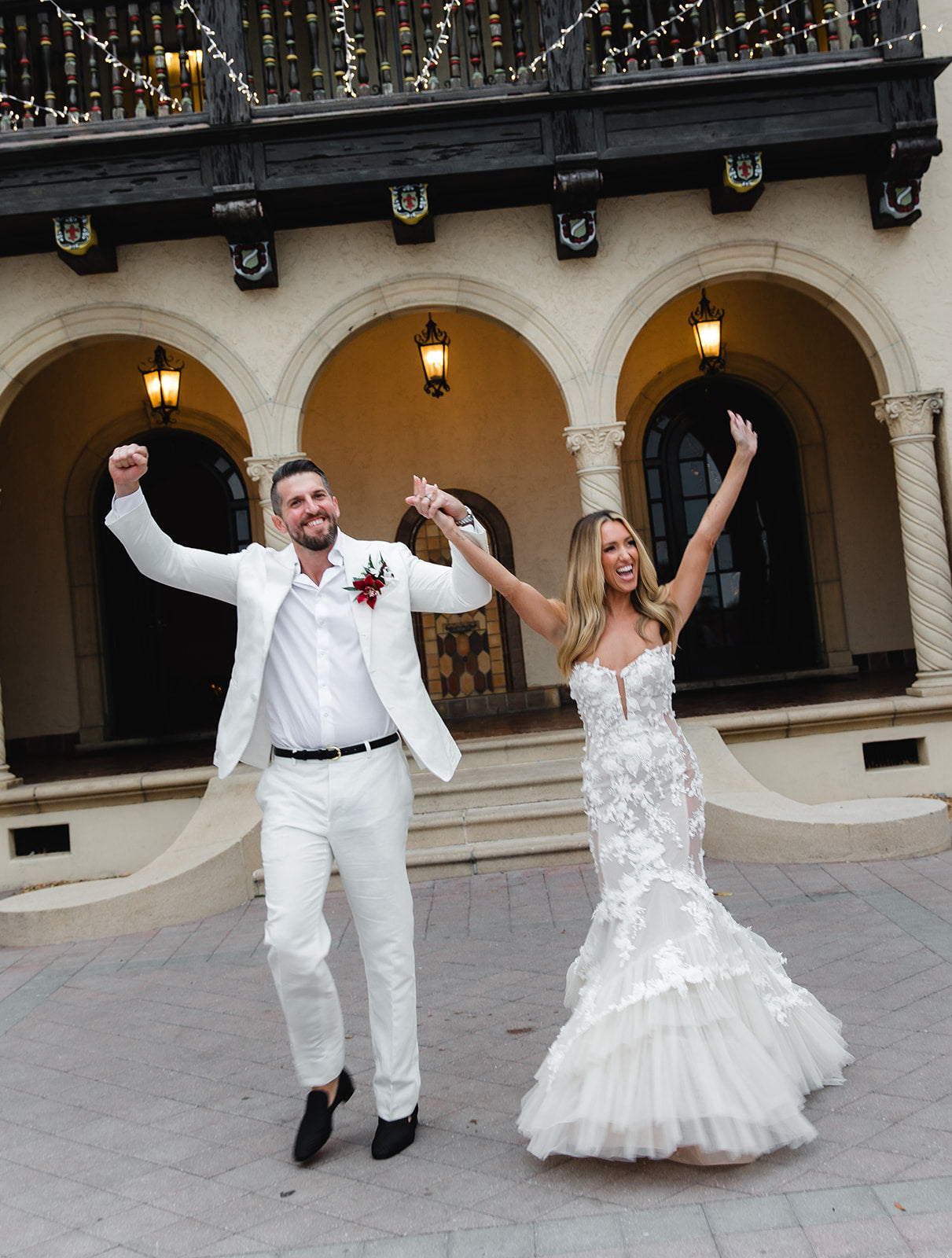 GL Bride Summer Fox and husband in wedding attire celebrating in front of a historic building. Bride Summer Fox in the lace Veronica dress by Galia Lahav 