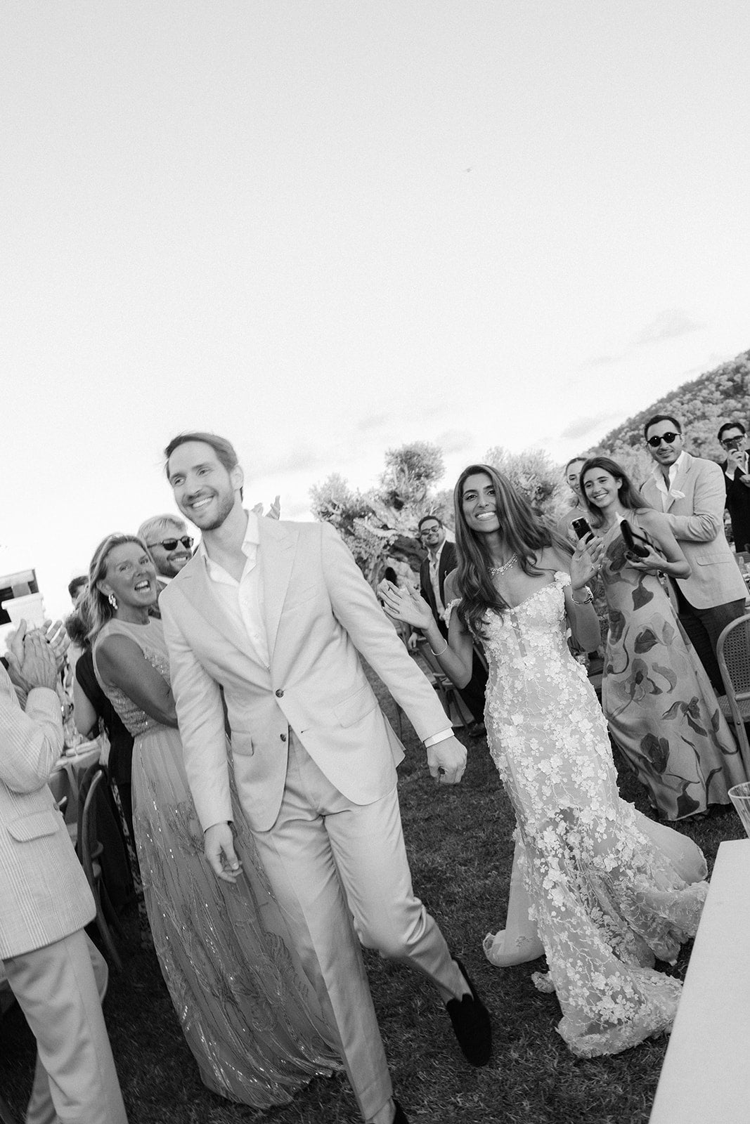 Black and white photo GL Bride Tara in our Maya gown and her husband walking through a crowd at an outdoor wedding.
