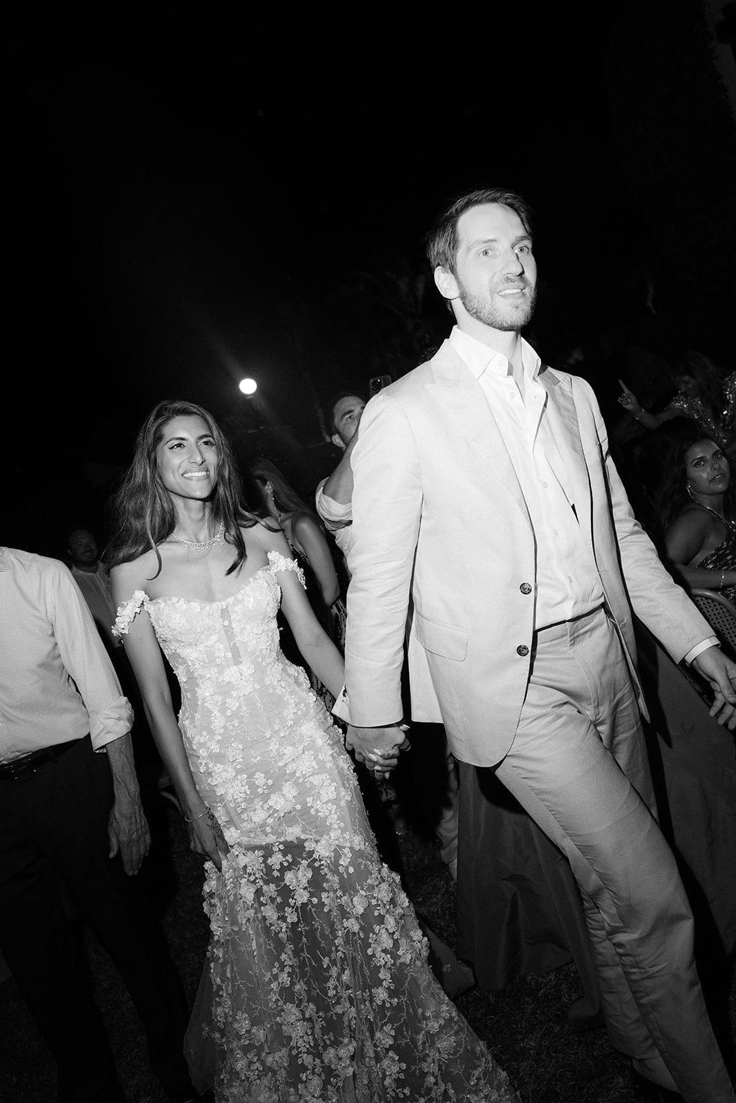 GL Bride Tara in our Maya gown and her husband walking together at their wedding event with a dark background