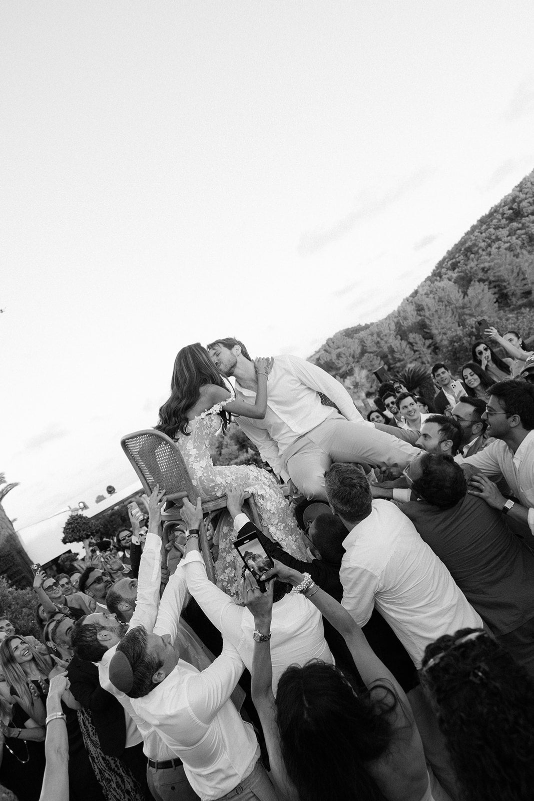 Black and white photo of a crowd lifting GL Bride Tara in our Maya gown and groom in chairs.
