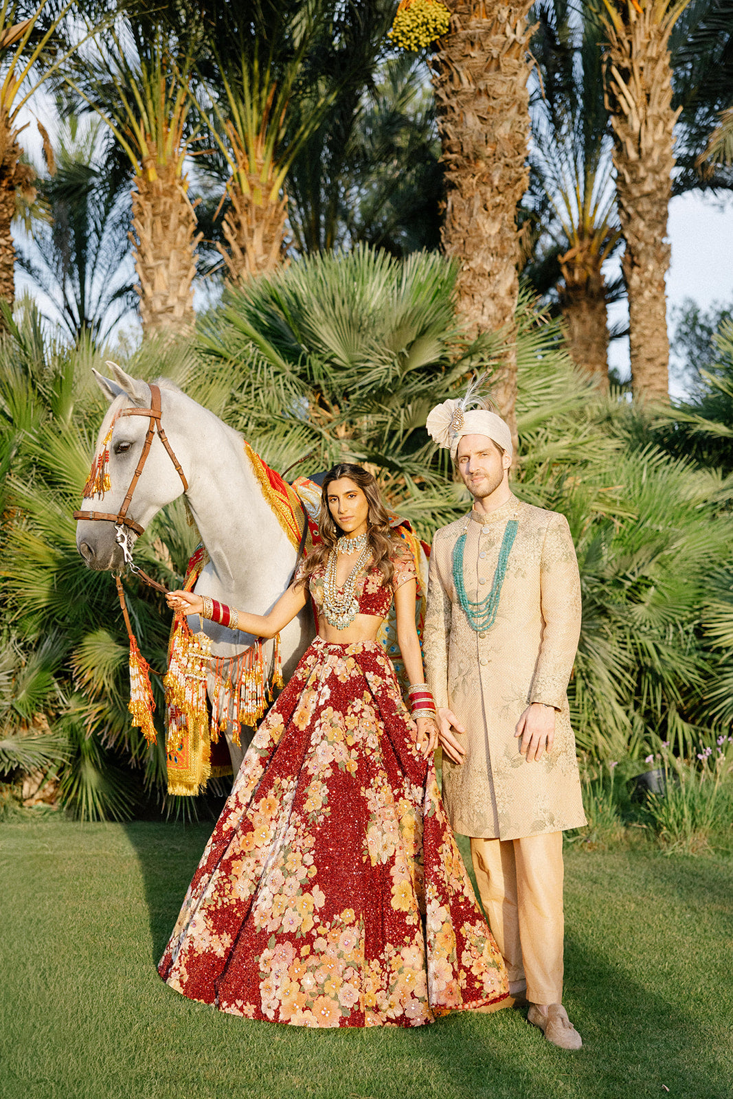 GL Bride Tara and her husband in traditional attire standing next to a horse with palm trees in the background