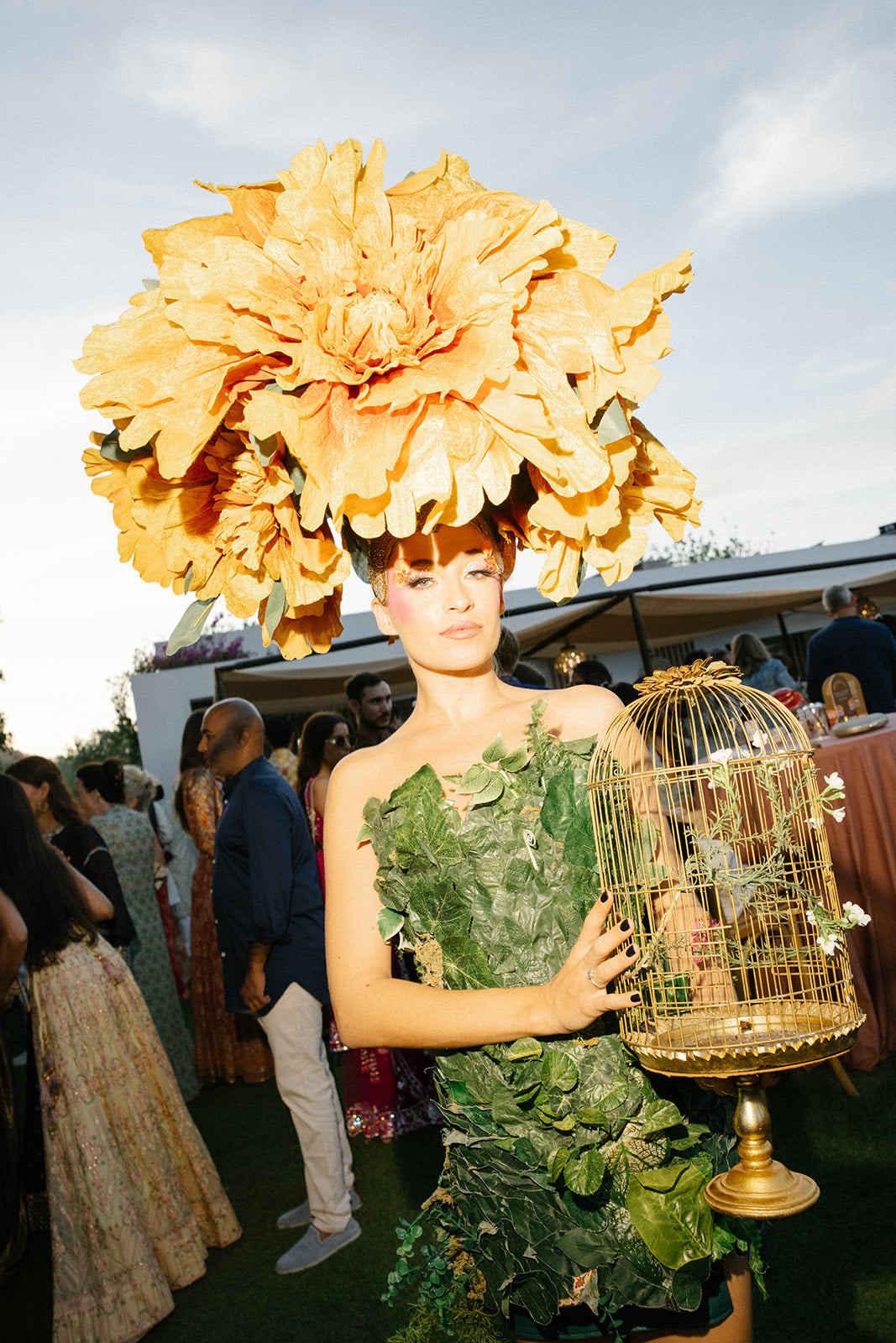 Woman in a green leafy dress holding a gold birdcage with a large yellow flower headpiece.