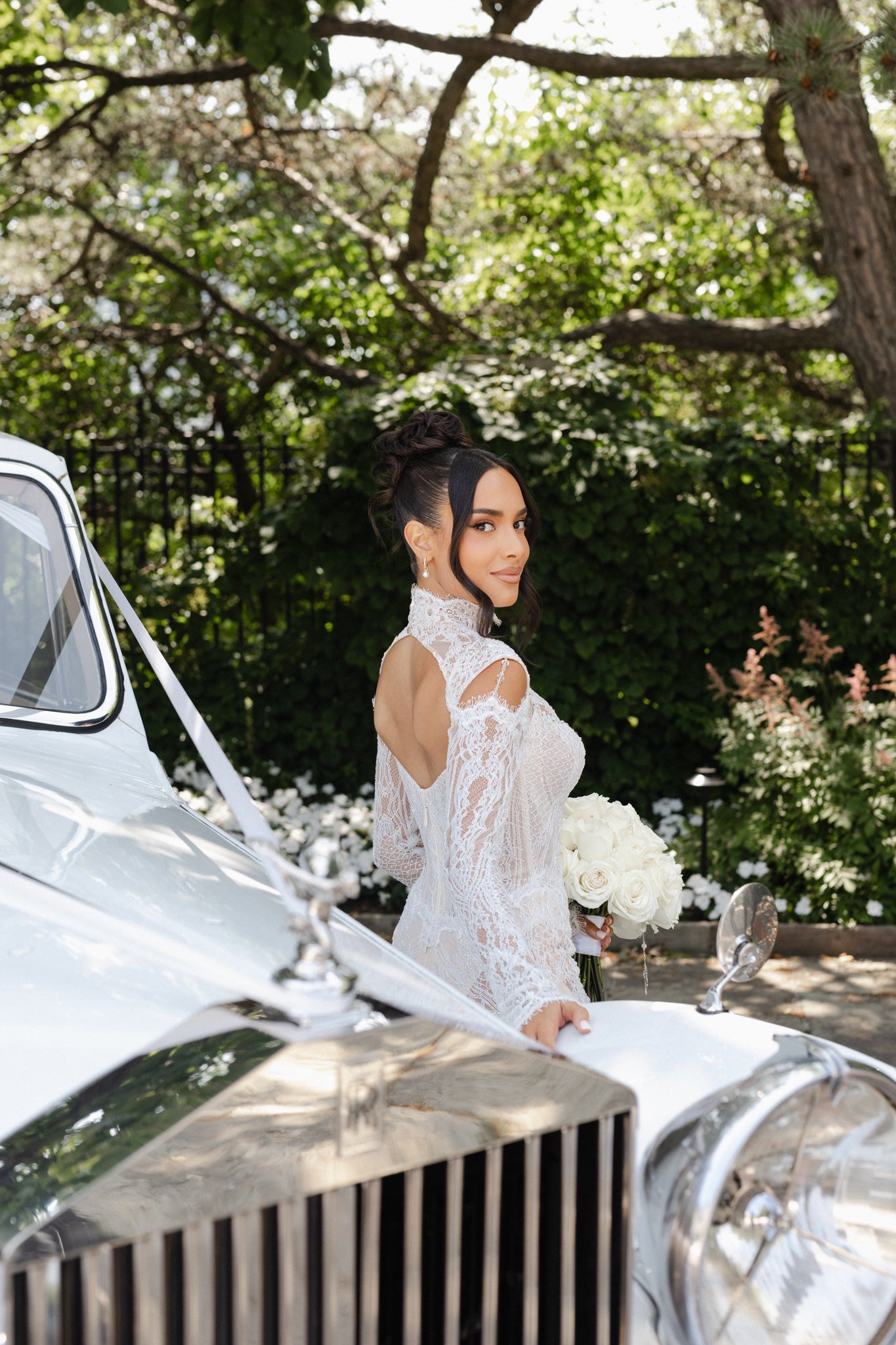 Woman in a white dress standing next to a vintage car with greenery in the background