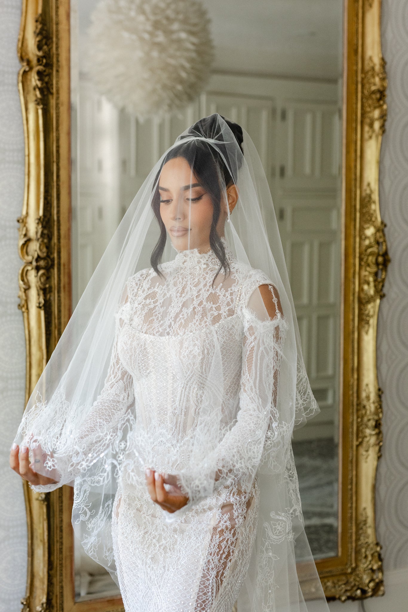 Woman in a white lace wedding dress with a veil standing in front of a large gold-framed mirror.