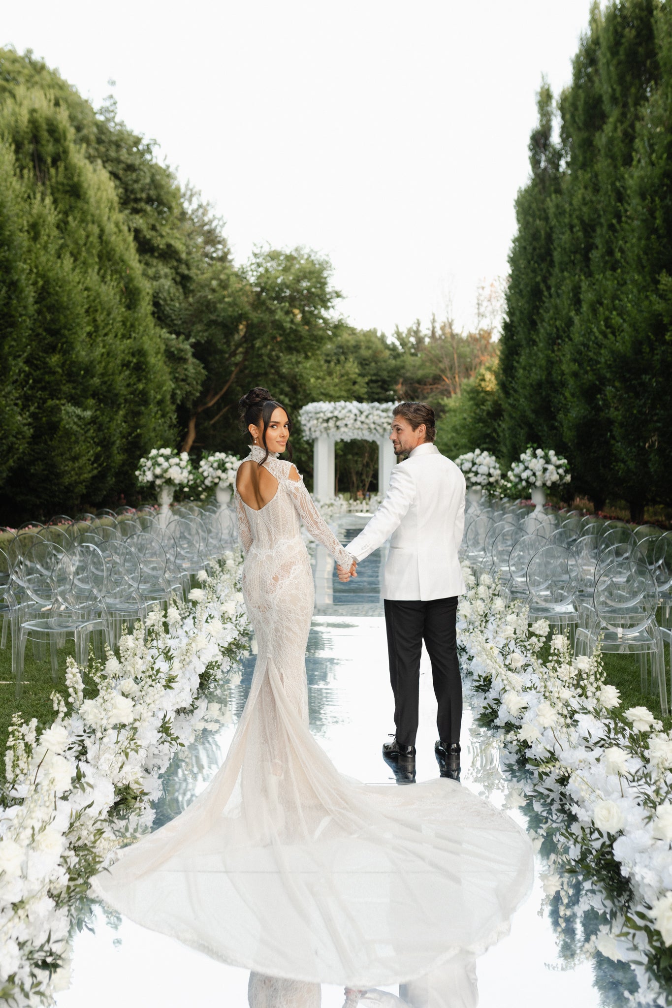 Couple holding hands in a garden with white floral arrangements and a white archway.