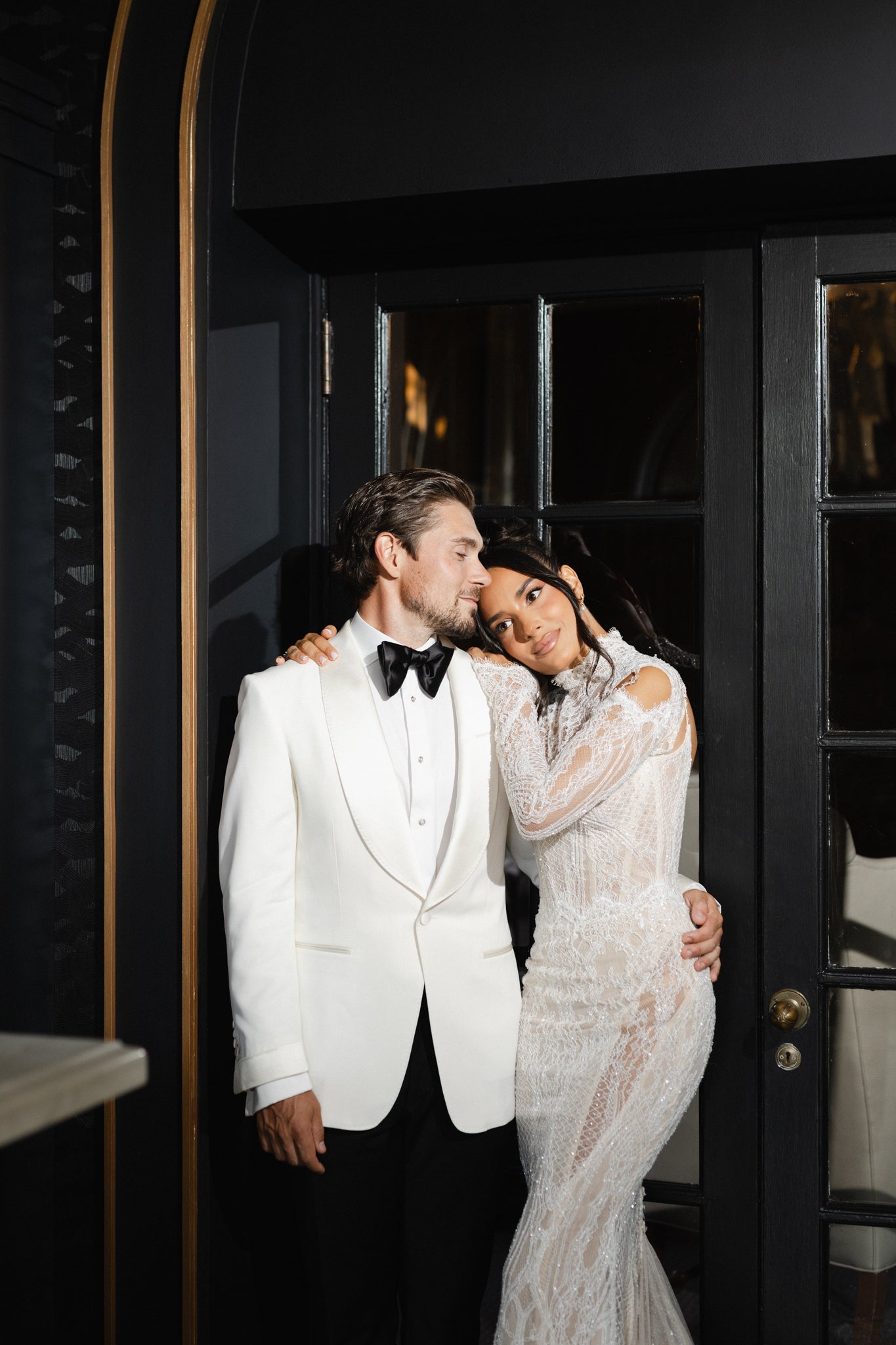 Man in a white tuxedo and woman in a white lace dress embracing in front of a black door.