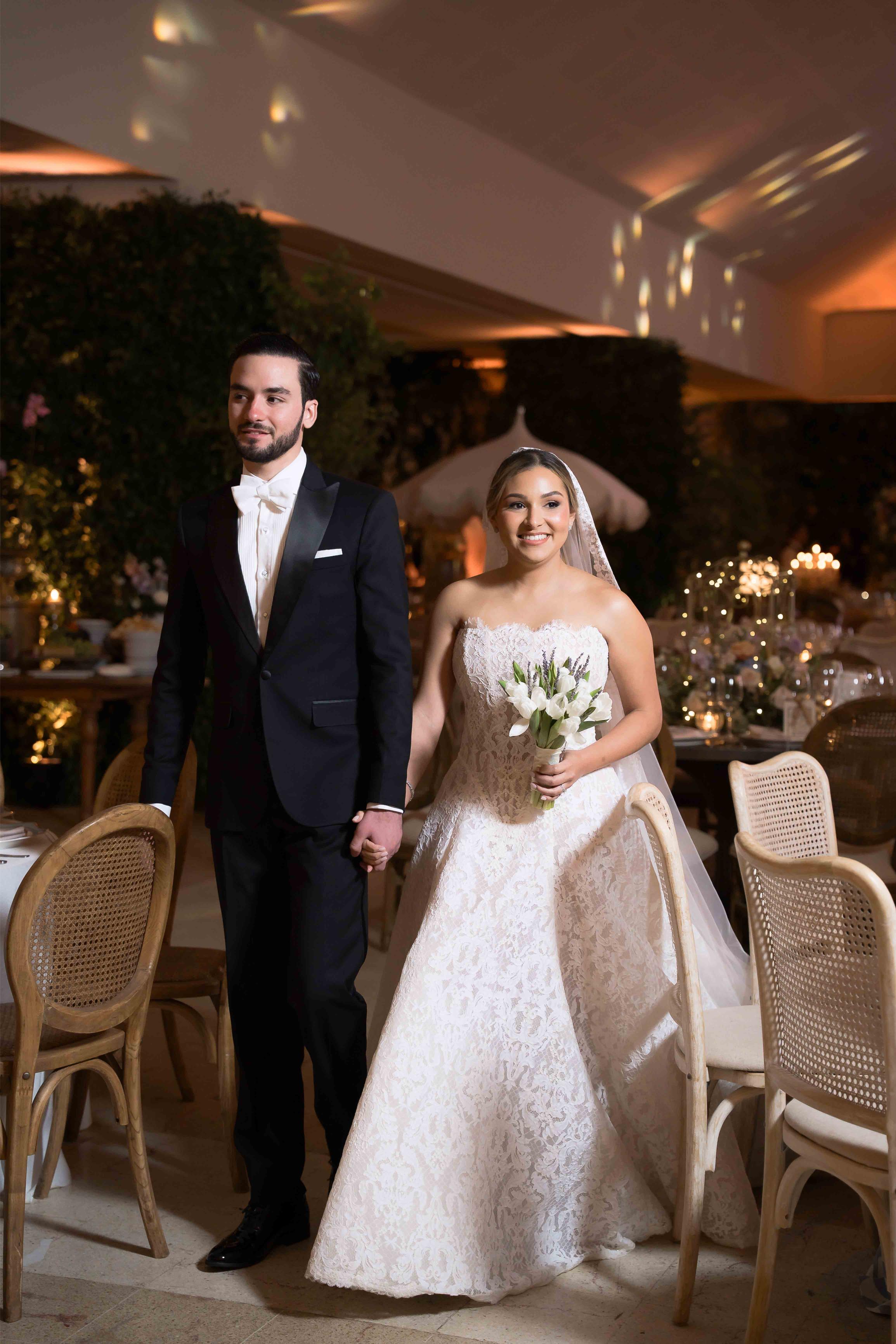 GL Bride Yamilca wearing our Splendid and her husband in wedding attire standing in a decorated indoor setting with tables and chairs.