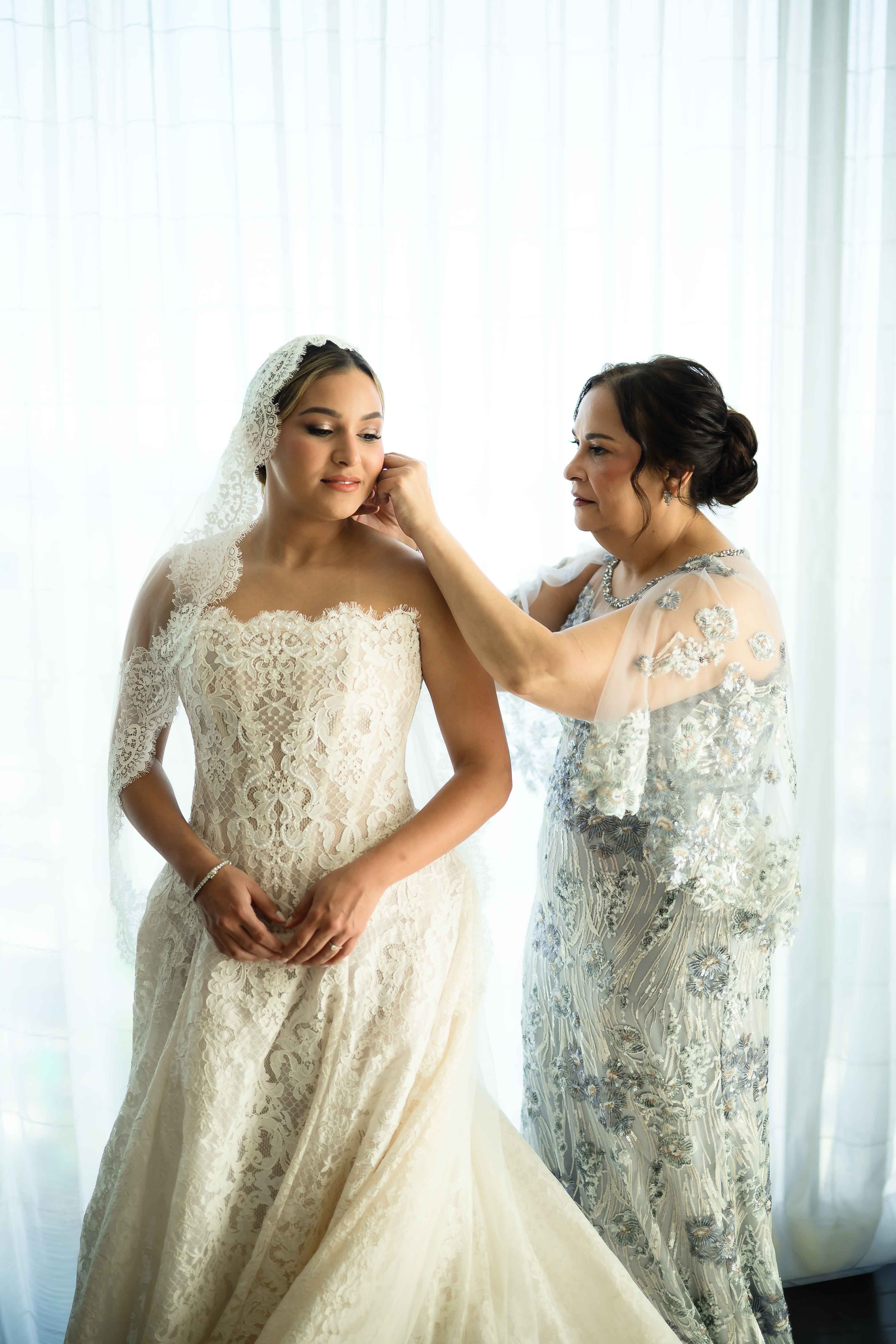GL Bride Yamilca wearing our Splendid, being help with her veil by her mother against a white curtain background