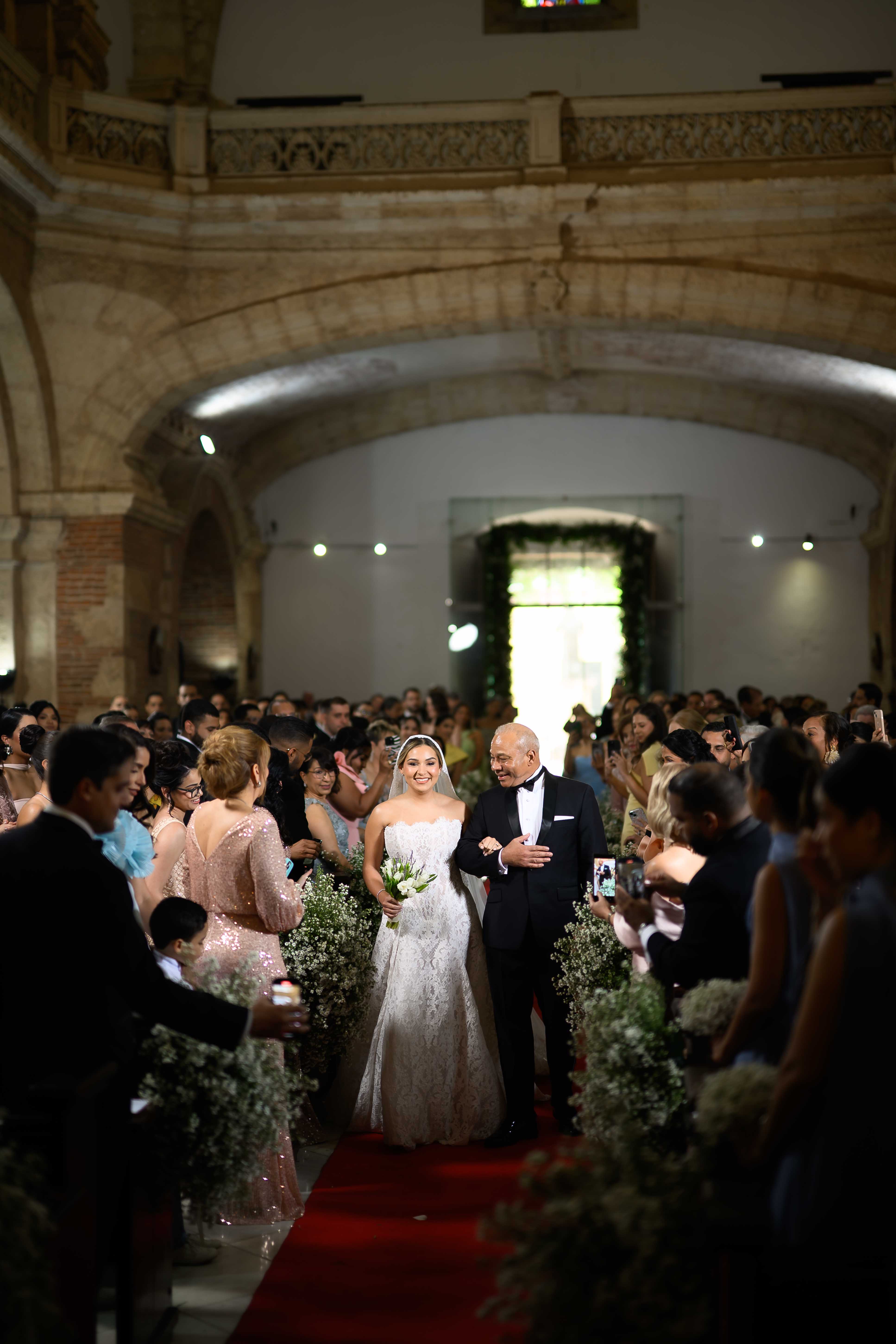 Wedding ceremony in a grand hall with guests and GL Bride Yamilca wearing our Splendid, and her father walking down the aisle.