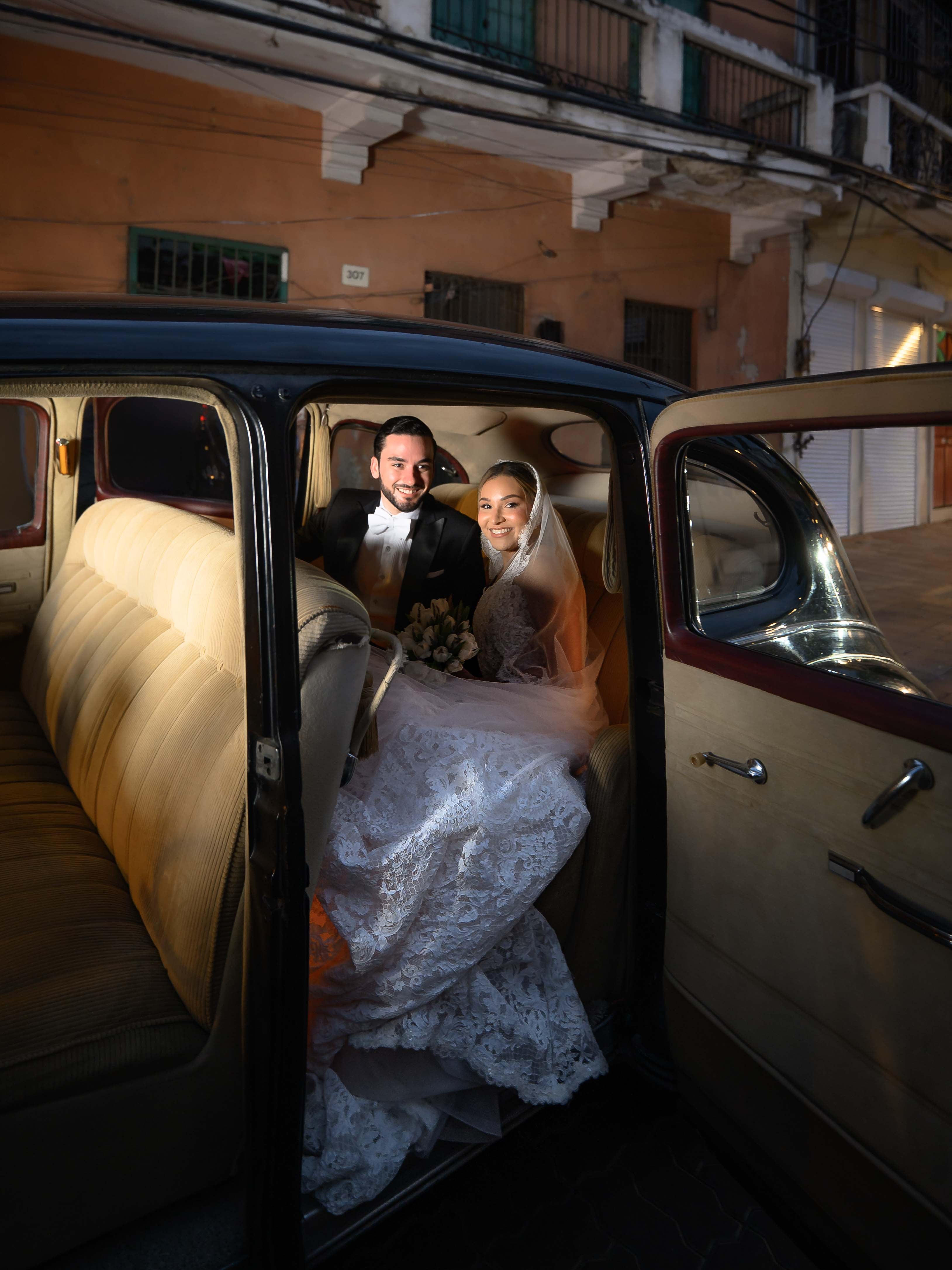 GL Bride Yamilca wearing our Splendid with her husband, sitting inside an open car door of a vintage vehicle.