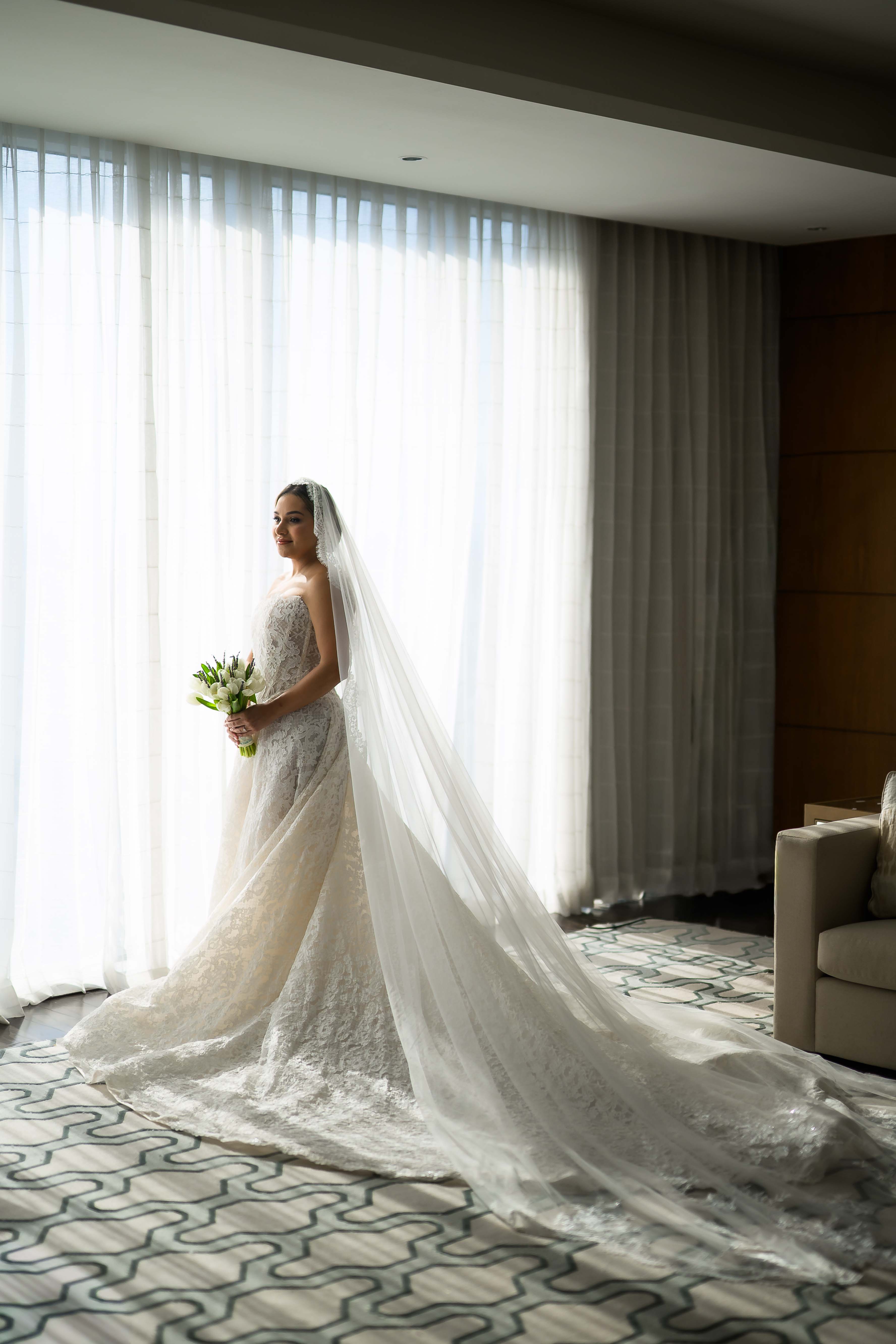 GL Bride Yamilca wearing our Splendid  with a long veil standing in a room with large windows.