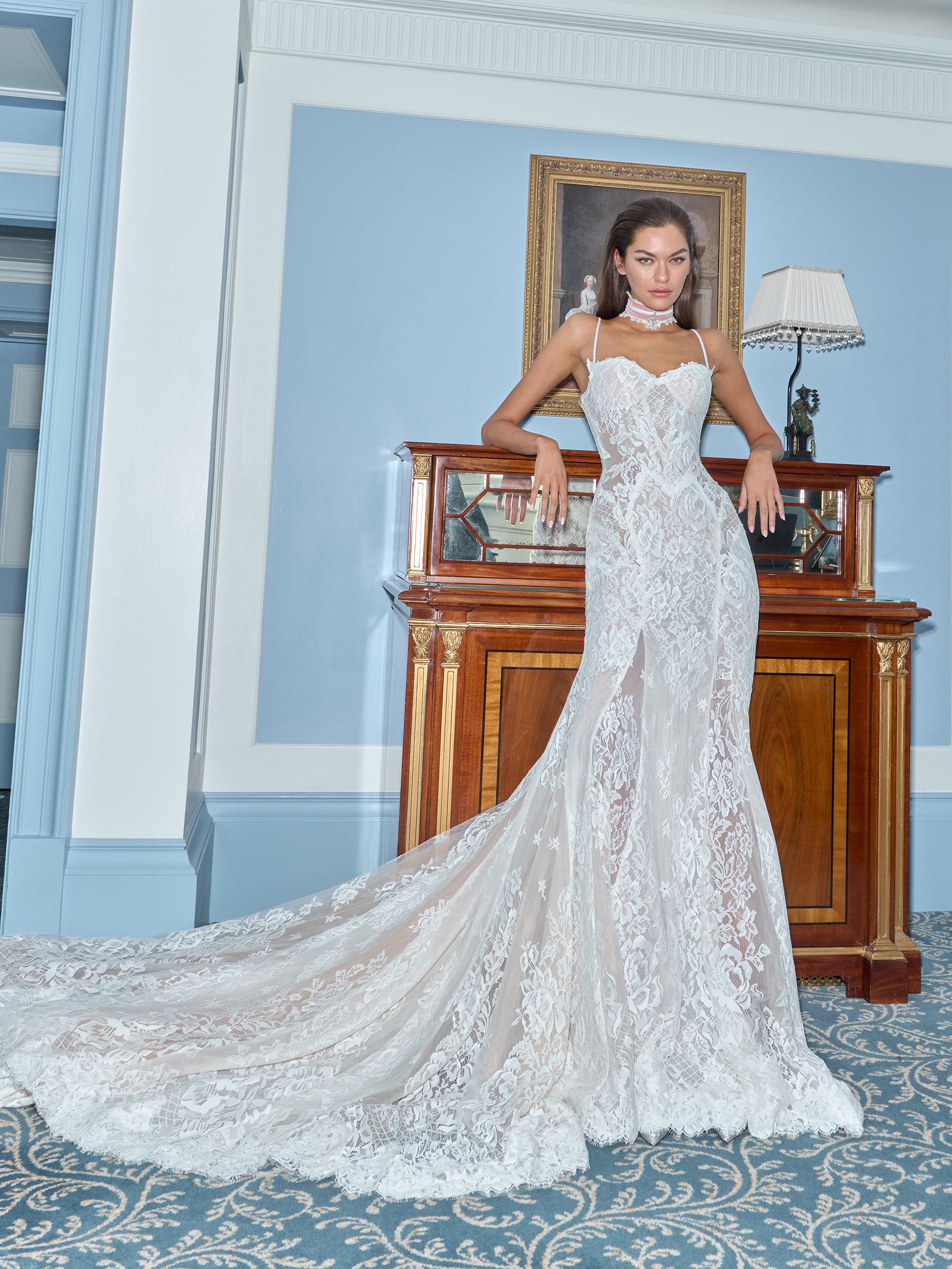 Woman in a white lace wedding dress standing in a room with a wooden fireplace and lamp.