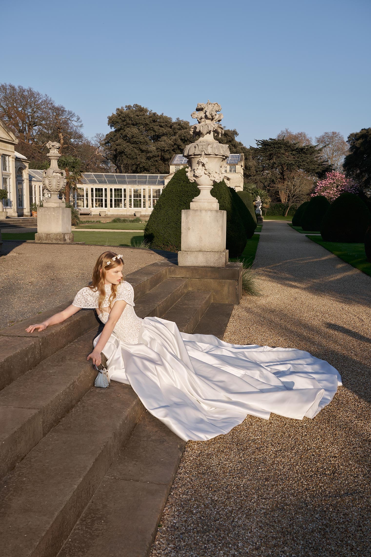 Woman in a white dress sitting on stone steps with classical statues in a garden setting