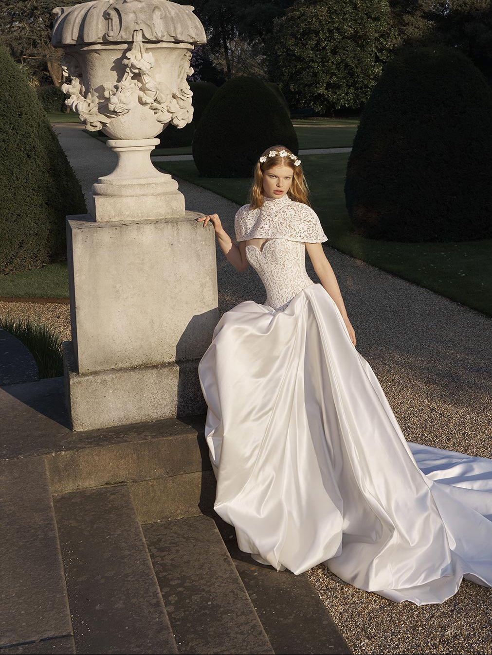 Woman in a white wedding dress standing next to a stone urn in a garden setting