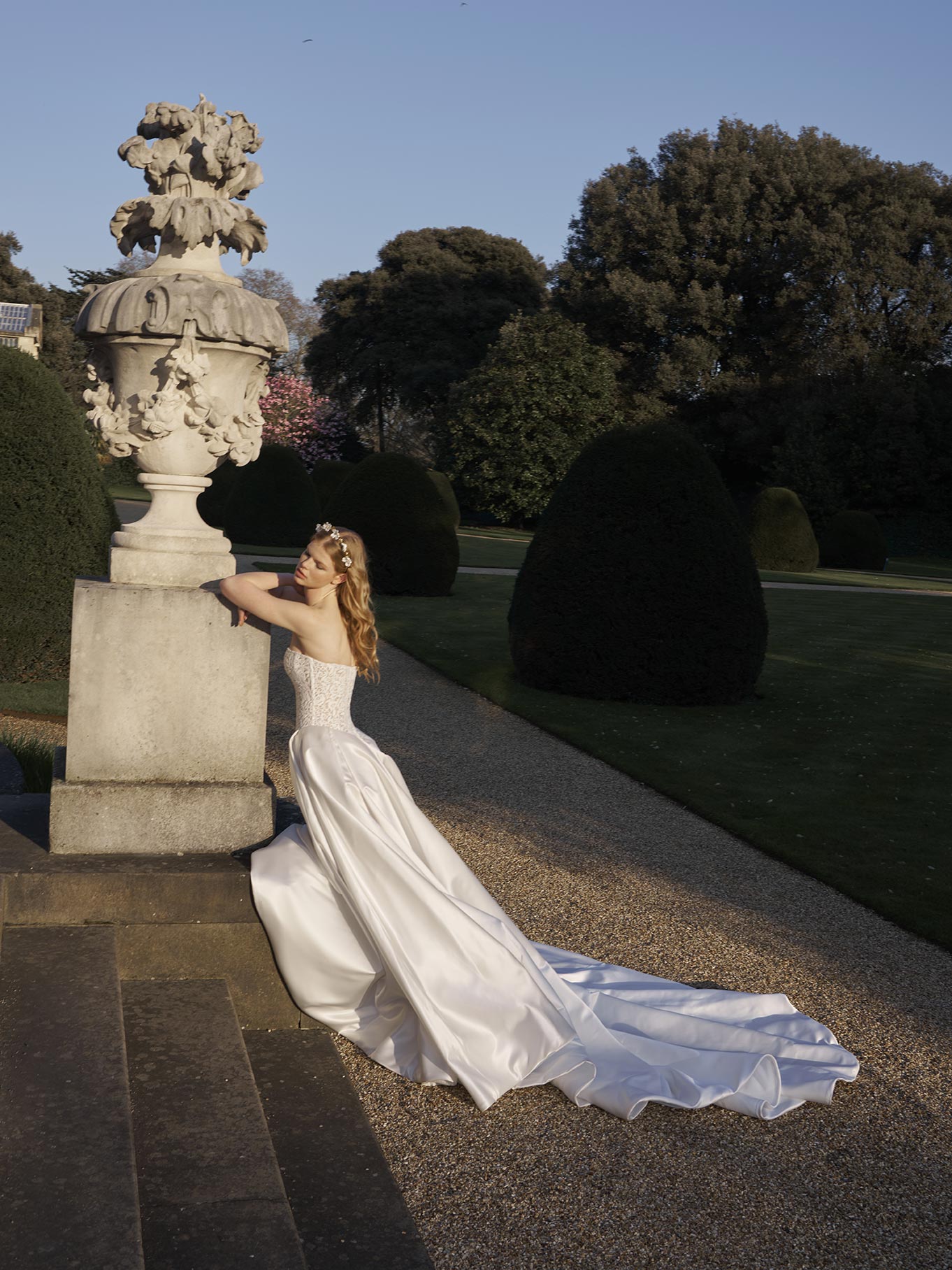 Woman in a white dress standing on stone steps with classical sculpture in a garden setting