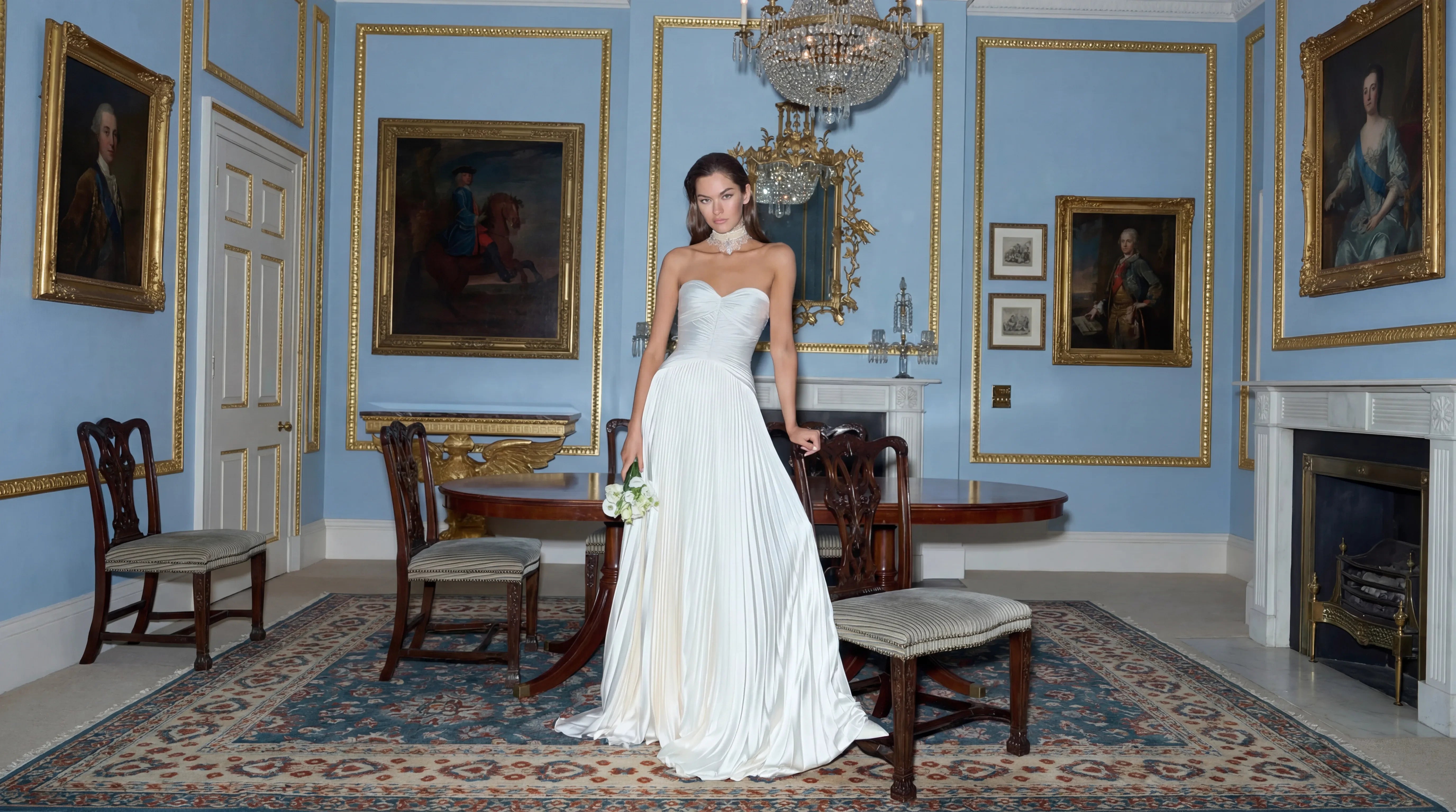 Woman in a white dress standing in an elegant room with blue walls, portraits, and a chandelier.