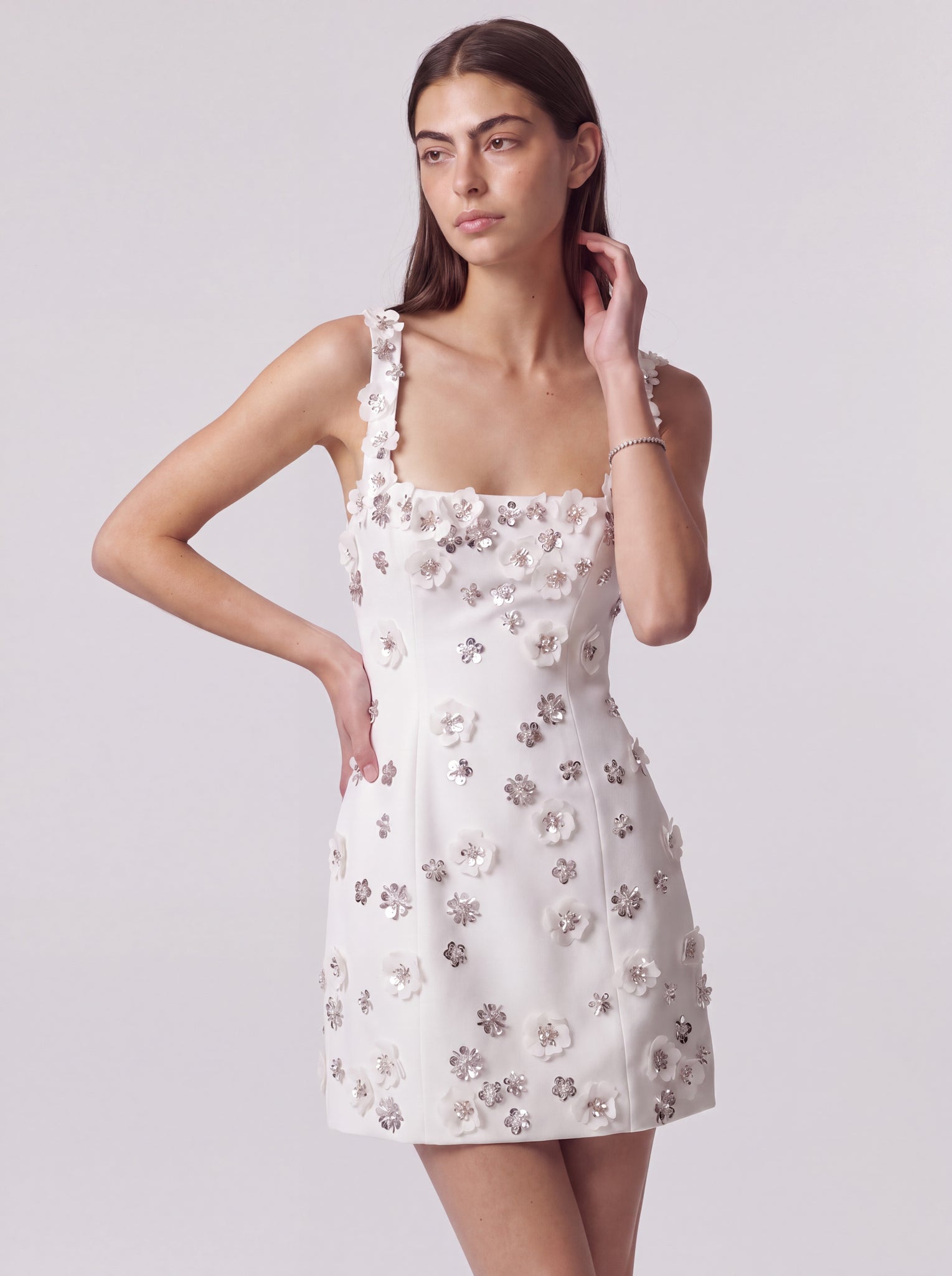A woman modeling a sleeveless white mini dress with 3D floral appliques in white and metallic silver, set against a plain light gray studio background.