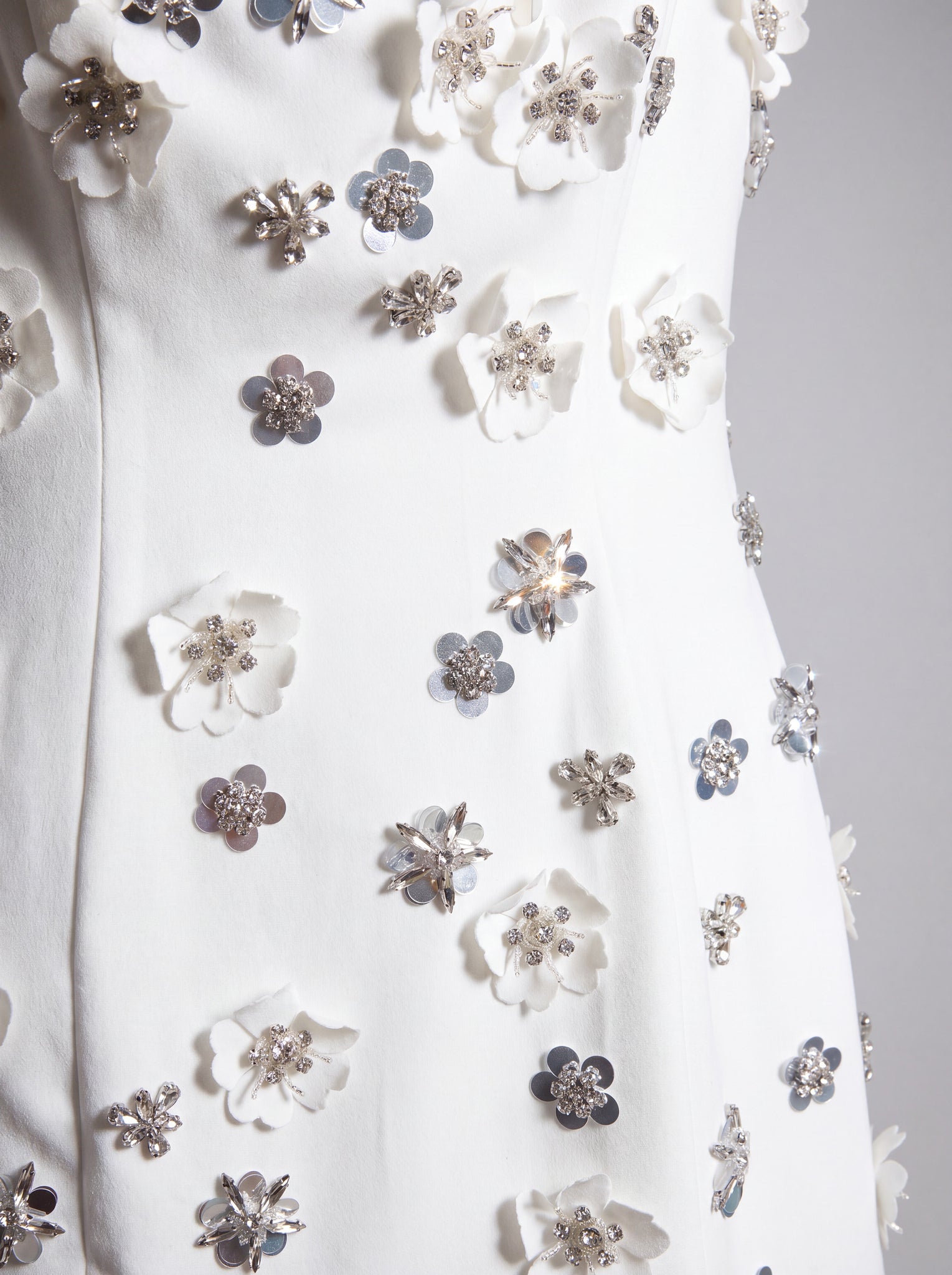 Close-up of a white garment featuring 3D floral appliqués in white and silver with rhinestone centers, displayed on a mannequin or dress form against a light gray background.