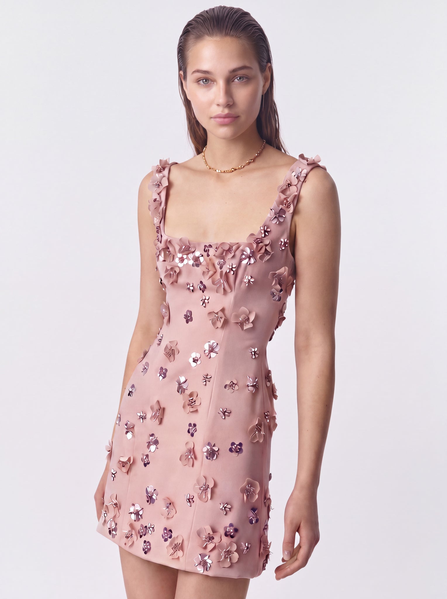 A model wearing a dusty pink sleeveless mini dress decorated with 3D floral appliqués and sequins, set against a plain light gray studio background.