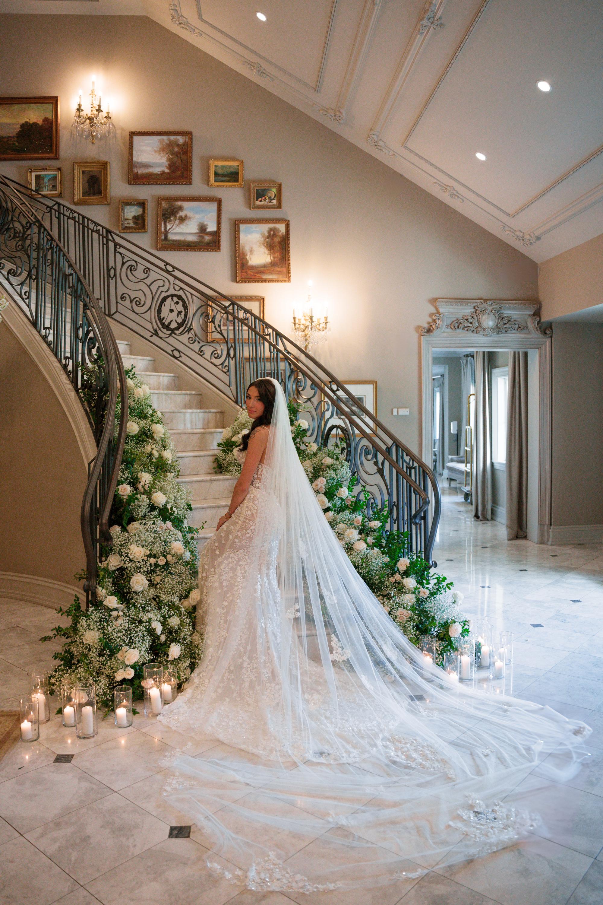 GL Bride Jessica in our Catalina gown standing on a decorated staircase with floral arrangements and candles.