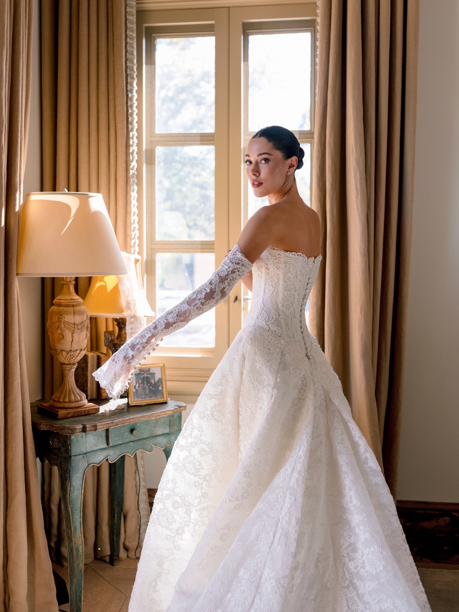 Woman in a white wedding dress standing in a room with curtains and a lamp.
