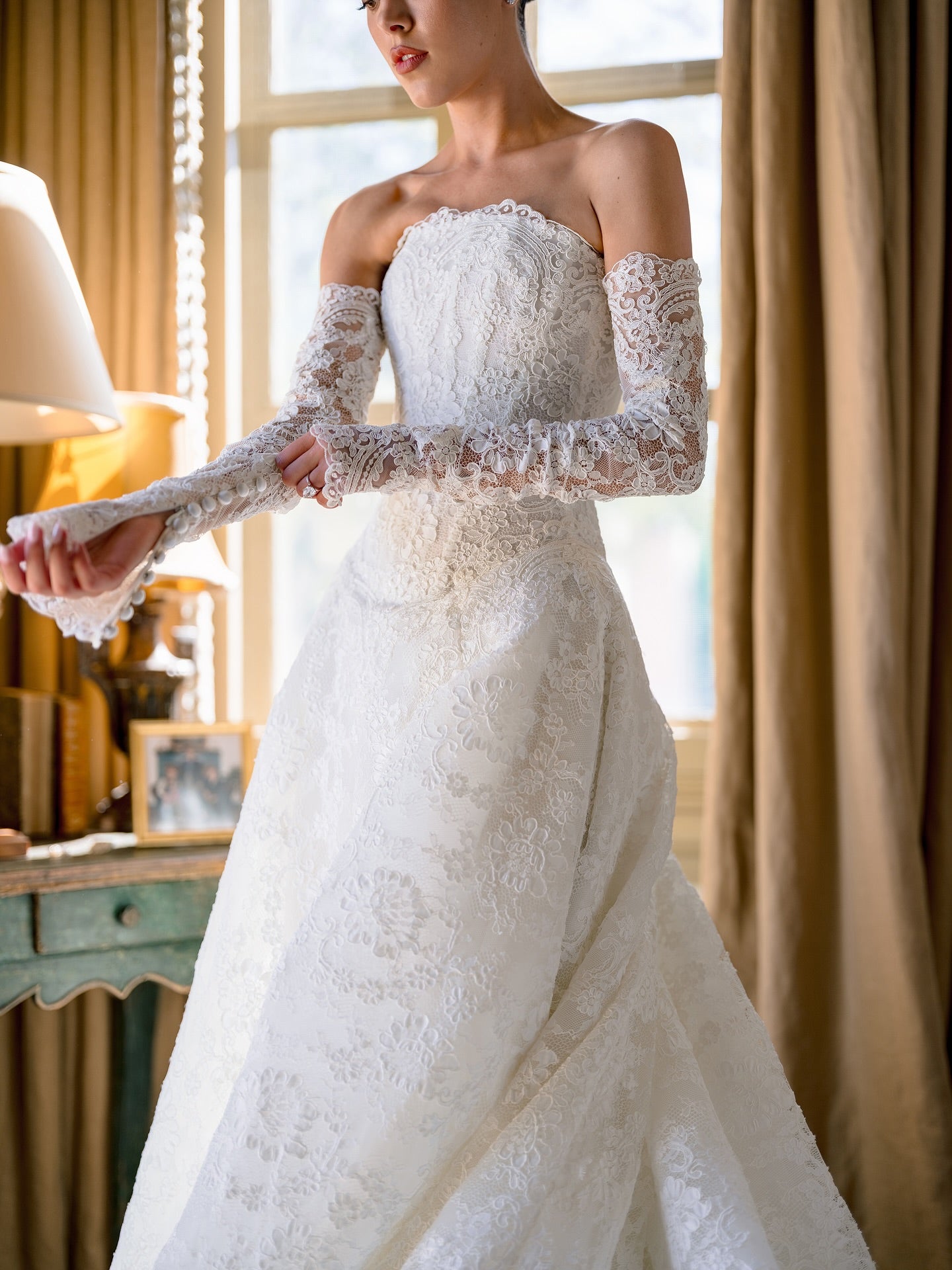 Woman wearing a white lace wedding dress with long sleeves in a room with curtains and a lamp.