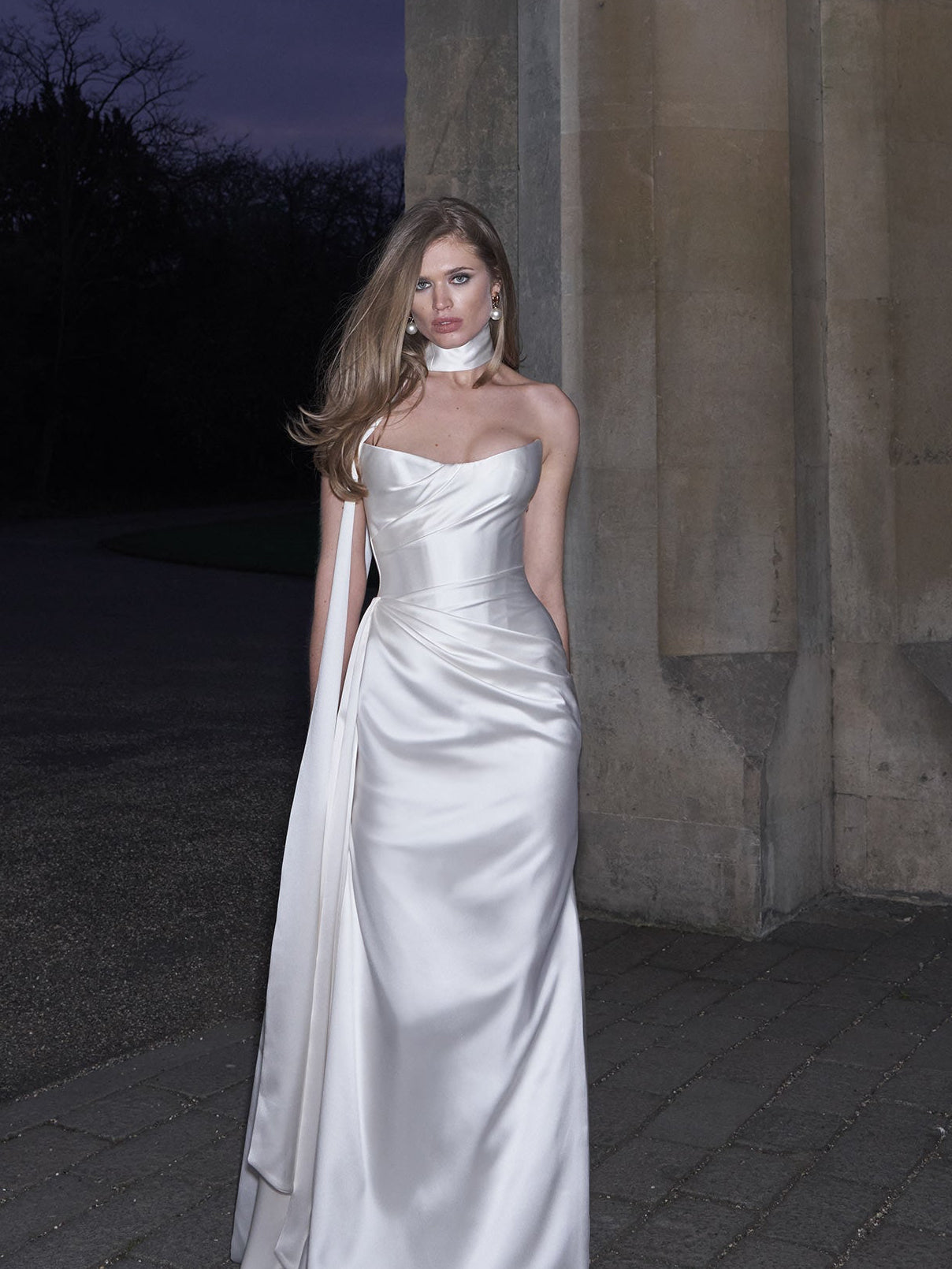 Woman in a white strapless gown standing against a stone wall at dusk.